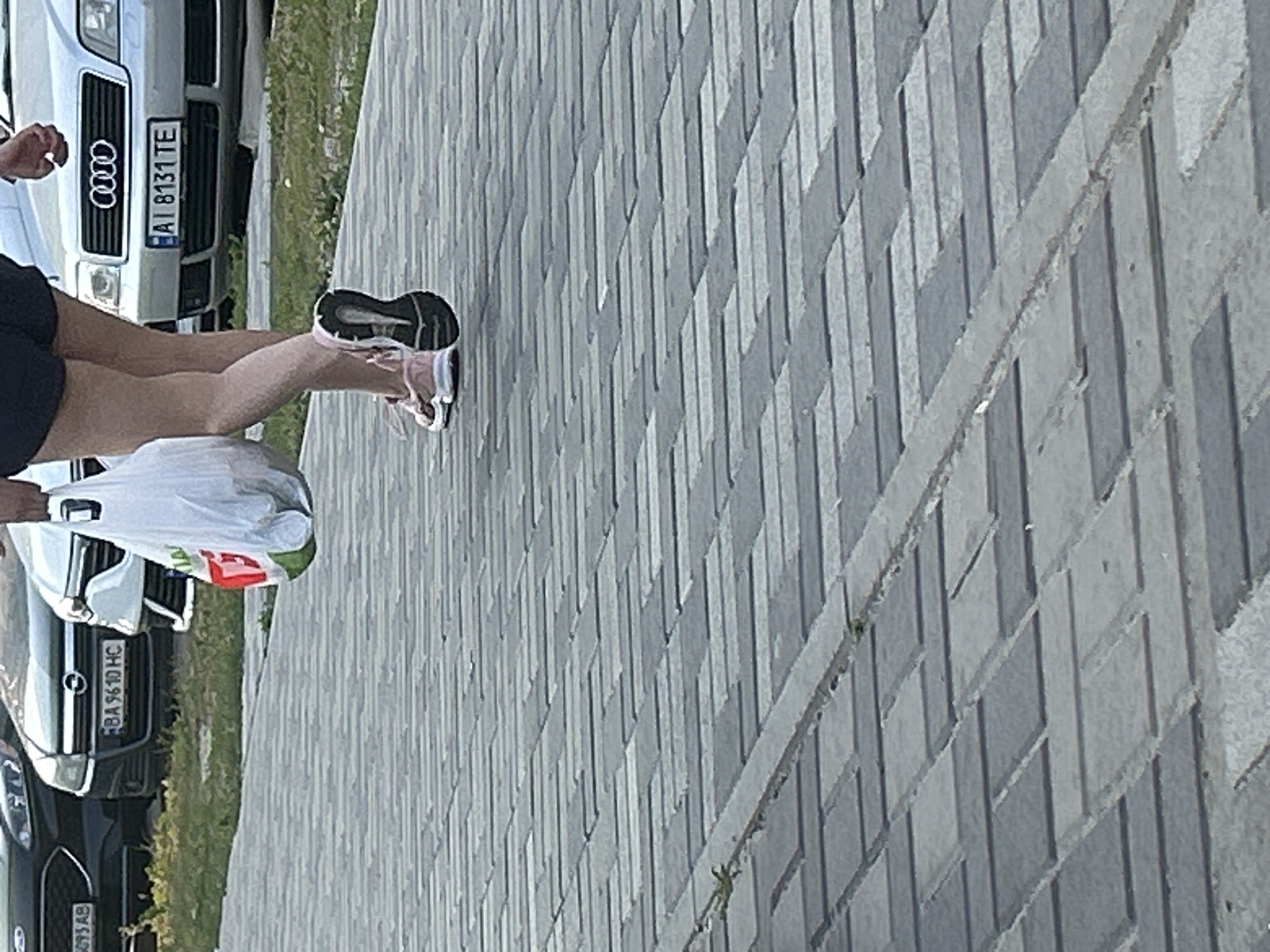 The image shows a person's legs and torso as they walk on a paved sidewalk. The person is carrying a white plastic bag in their right hand. They are wearing dark shorts and pink and white athletic shoes. In the background, several cars are parked, including a silver Audi with license plate AI 8131 TE. The scene appears to be outdoors, likely on a sunny day, in the city of Brovary, Ukraine. The paving stones of the sidewalk are made of rectangular concrete blocks arranged in a pattern.