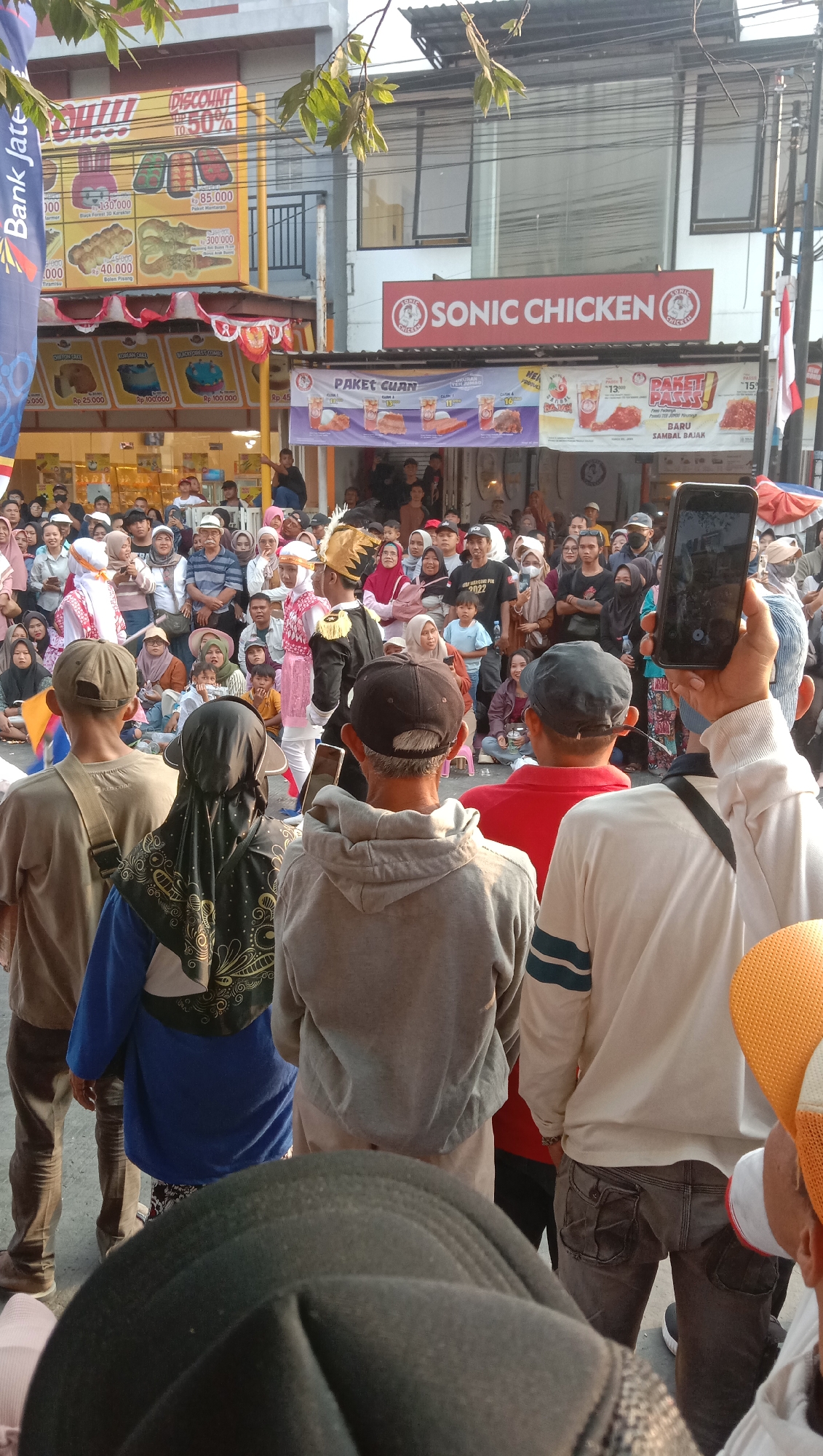 In the city of Jatisawit, Indonesia, a crowd gathers in front of a Sonic Chicken establishment. The scene appears to be a lively event, possibly a festival or promotion, with people of all ages present. Some individuals are dressed in traditional attire, while others are in casual clothing. The Sonic Chicken store has banners displaying food promotions, including "Paket Cuan" and "Paket Puass," offering various meal deals. A performer in a black and gold uniform, with a golden headdress, stands out in the center, interacting with the crowd. A person in the foreground is holding up a smartphone, seemingly recording or photographing the event. The environment is urban, with buildings and streetlights visible in the background. The overall atmosphere suggests a community gathering and a celebration of local culture and commerce.