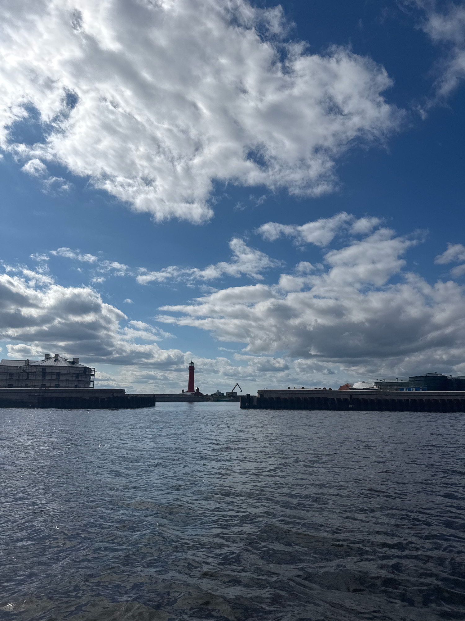 The image depicts a view of the Kronstadt seaport in Russia on a partly cloudy day. In the foreground, dark blue water with ripples stretches across the frame. In the middle ground, a concrete pier separates the water from the background. On the pier, a tall, red lighthouse stands prominently, with a building to its left and other structures and industrial equipment, including a crane, visible on the pier to the right. The sky above is a bright blue, interspersed with dramatic, white and grey clouds. The overall scene captures a maritime industrial landscape.
