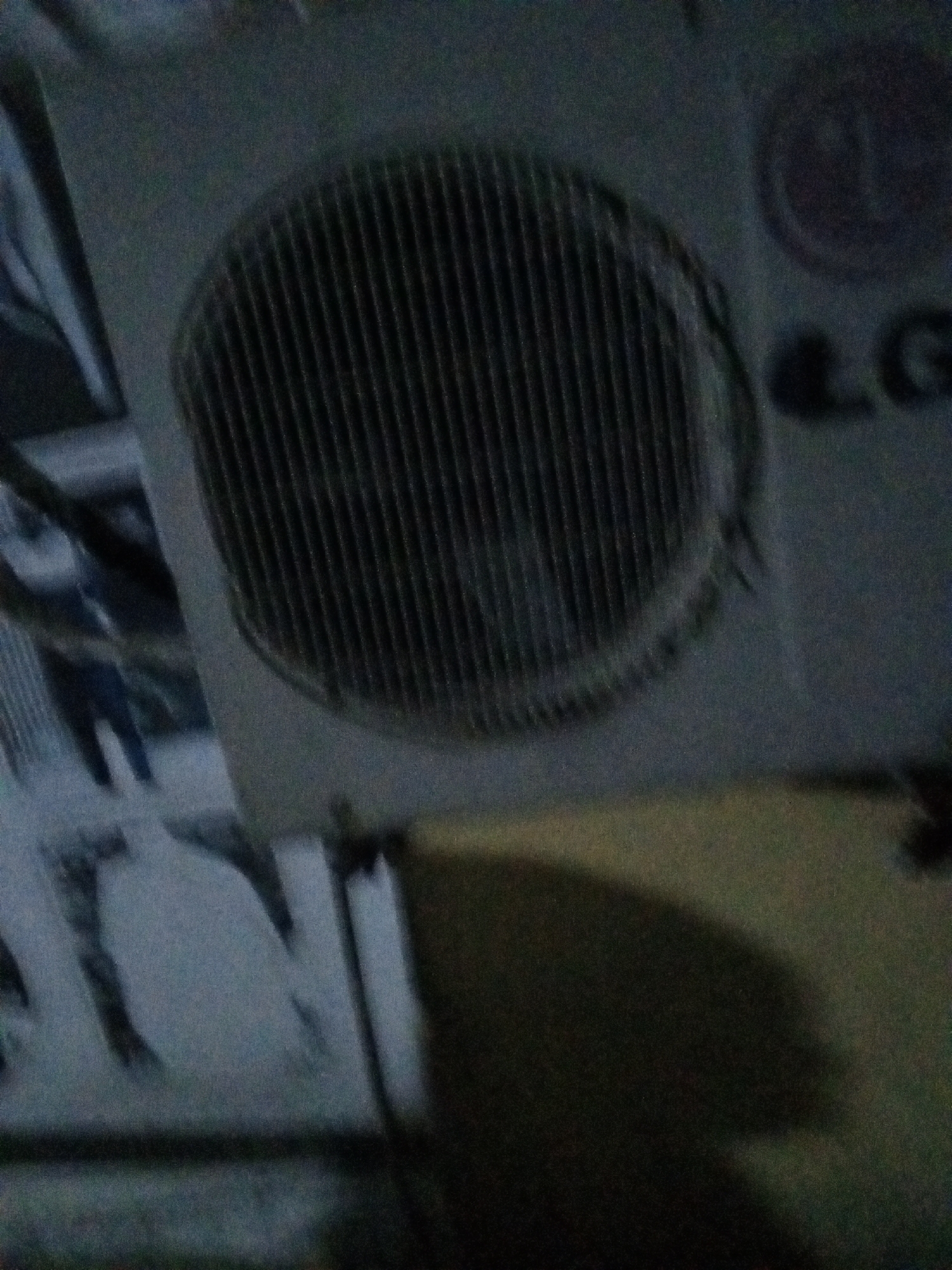 An LG window air conditioner is mounted in a wall in Ado, Nigeria. The exterior grill has a circular pattern, and the LG logo is visible on the right side of the unit. The wall is a light color, and a dark shadow is cast below the air conditioner. There is a window to the left of the AC unit.