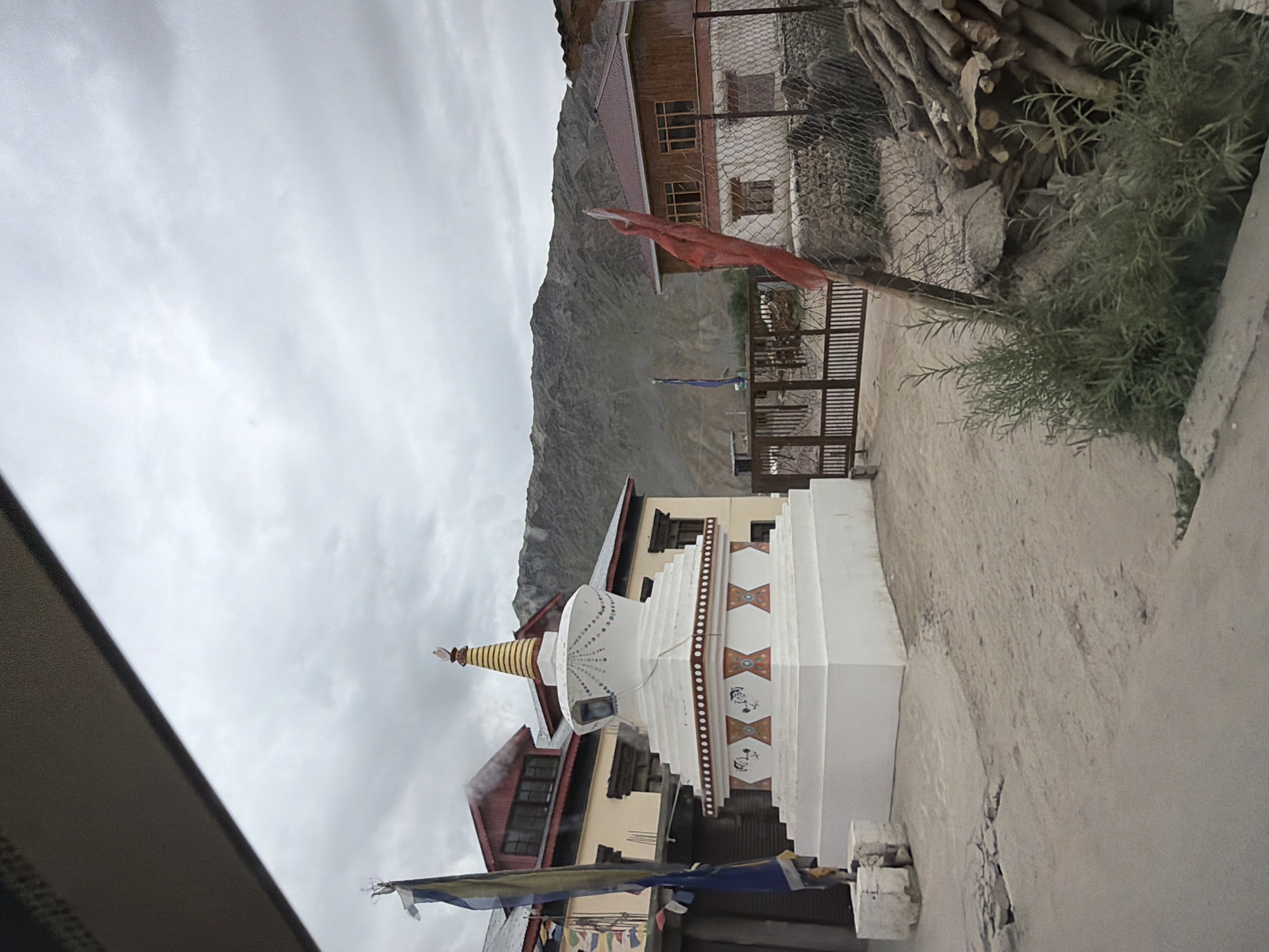 The image captures a scene in Padum, India, featuring a white stupa with decorative bandings and a conical, golden top. To the left of the stupa, a flagpole with prayer flags is visible, alongside a building with a red roof and traditional windows. A metal gate is positioned in the middle ground, leading to another building with a wooden exterior and a red roof. The background is dominated by a large, barren mountain under a cloudy sky. In the foreground, to the right, a pile of logs rests against a wire fence.