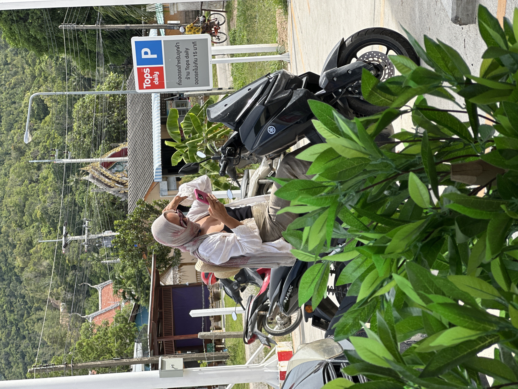 A person wearing a light-colored hijab and sunglasses is sitting on a black scooter, looking at their pink smartphone. They are wearing a white open-buttoned shirt over a dark tank top and khaki pants. A straw hat is placed on the back of the scooter. In the background, there is a street with a sign indicating "Tops daily" parking, a red and white striped bollard, other scooters, a distant person on a bicycle, and a lush green, tree-covered hillside. The scene appears to be in a tropical or sub-tropical urban environment.