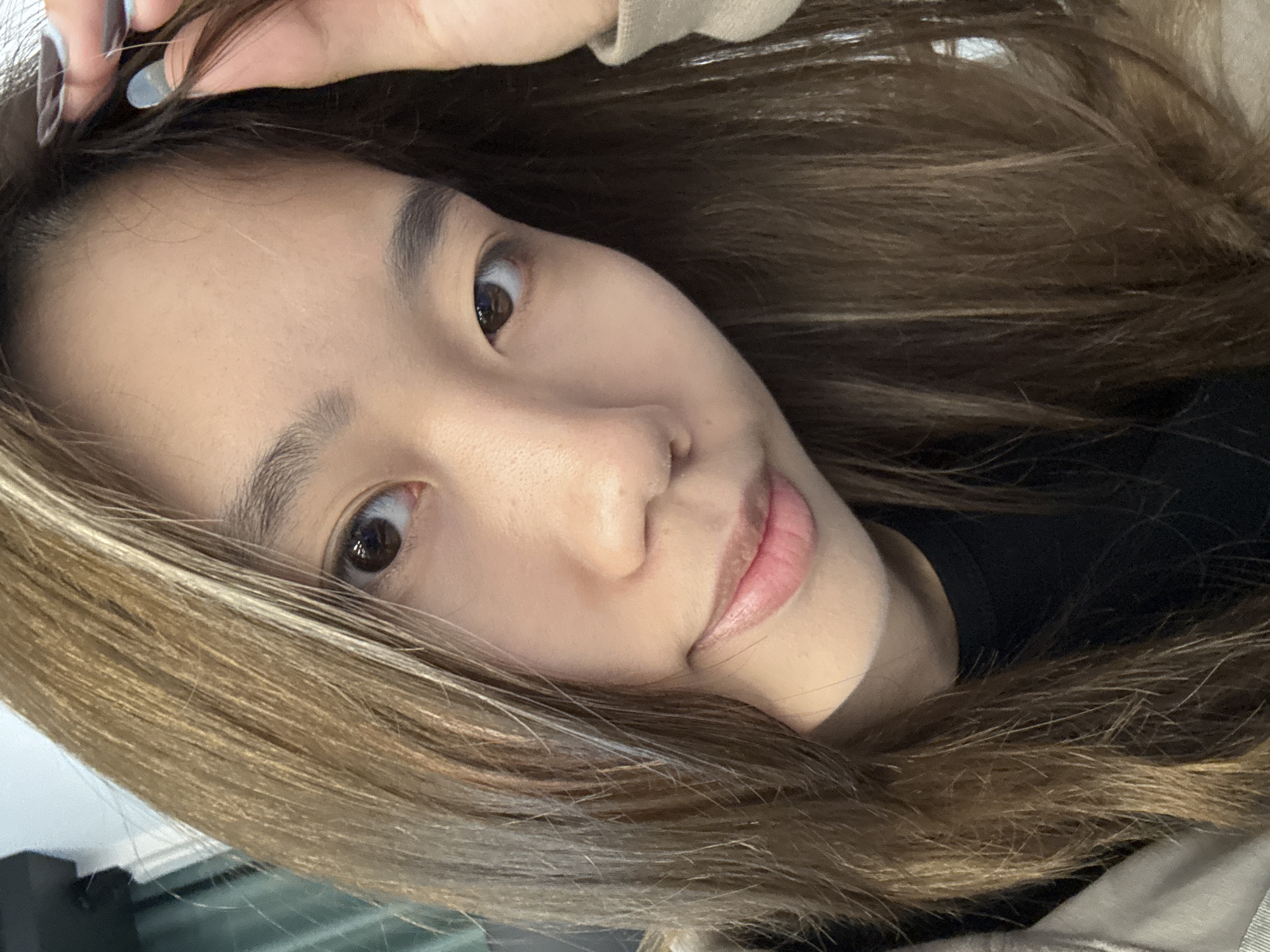 A close-up, slightly angled selfie of a young Asian woman with long, light brown hair. Her hair frames her face, and she has one hand near her head. She has brown eyes, fair skin, and pink lips. She is wearing a dark shirt. The background is blurry and indistinct, suggesting the location is an indoor space. There is no evidence that this picture was taken in Nan'an District, China.