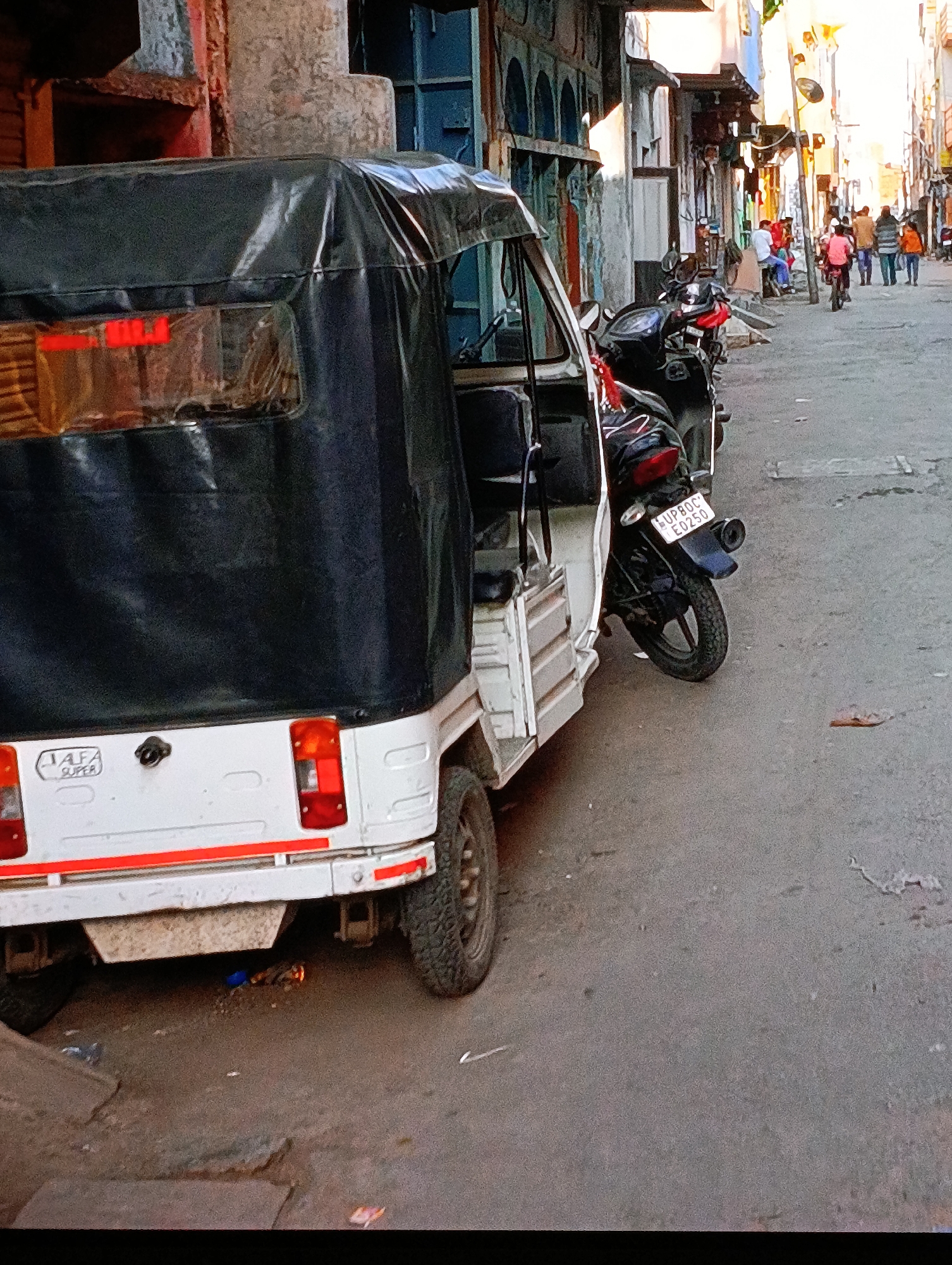 The image depicts a street scene in an unidentified city. In the foreground is a three-wheeled auto-rickshaw, white with a black canopy, parked on the side of the street. A motorcycle is parked next to it, with its license plate visible, reading "UP80C E0250". The street leads into the distance, where a group of people can be seen walking away. The buildings on either side of the street appear to be shops or residences, with a variety of architectural styles and colors. The overall impression is of a busy, everyday scene in an urban environment.