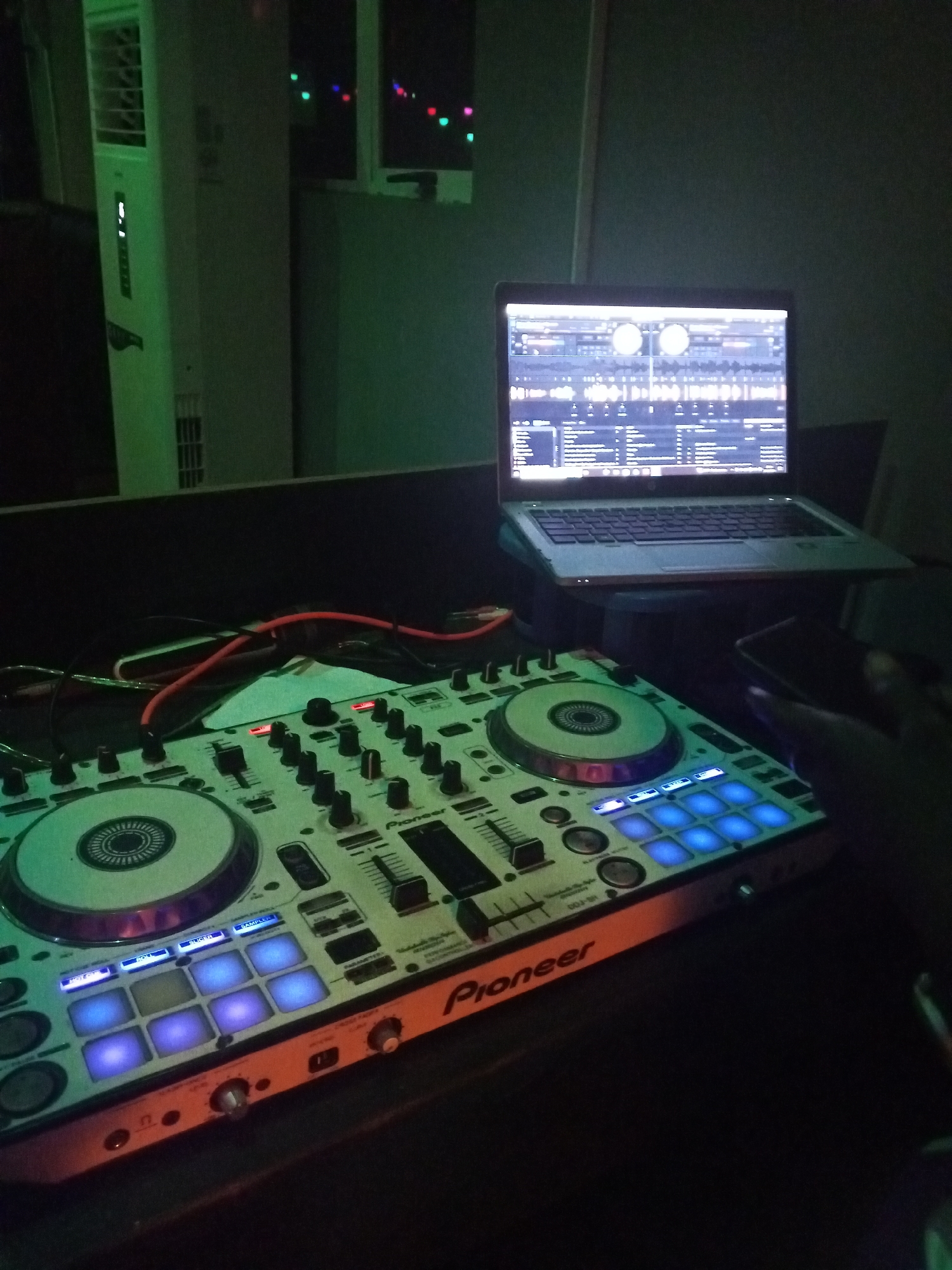 The image shows a DJ setup indoors, likely in Lokoja, Nigeria, based on the user's prompt. A Pioneer DJ controller is in the foreground with a laptop behind it displaying DJ software. Cables are connected to the controller. A person's hand holding a phone is partially visible to the right. In the background, there is an air conditioning unit and a window with lights outside. The room appears dimly lit with a green tint.