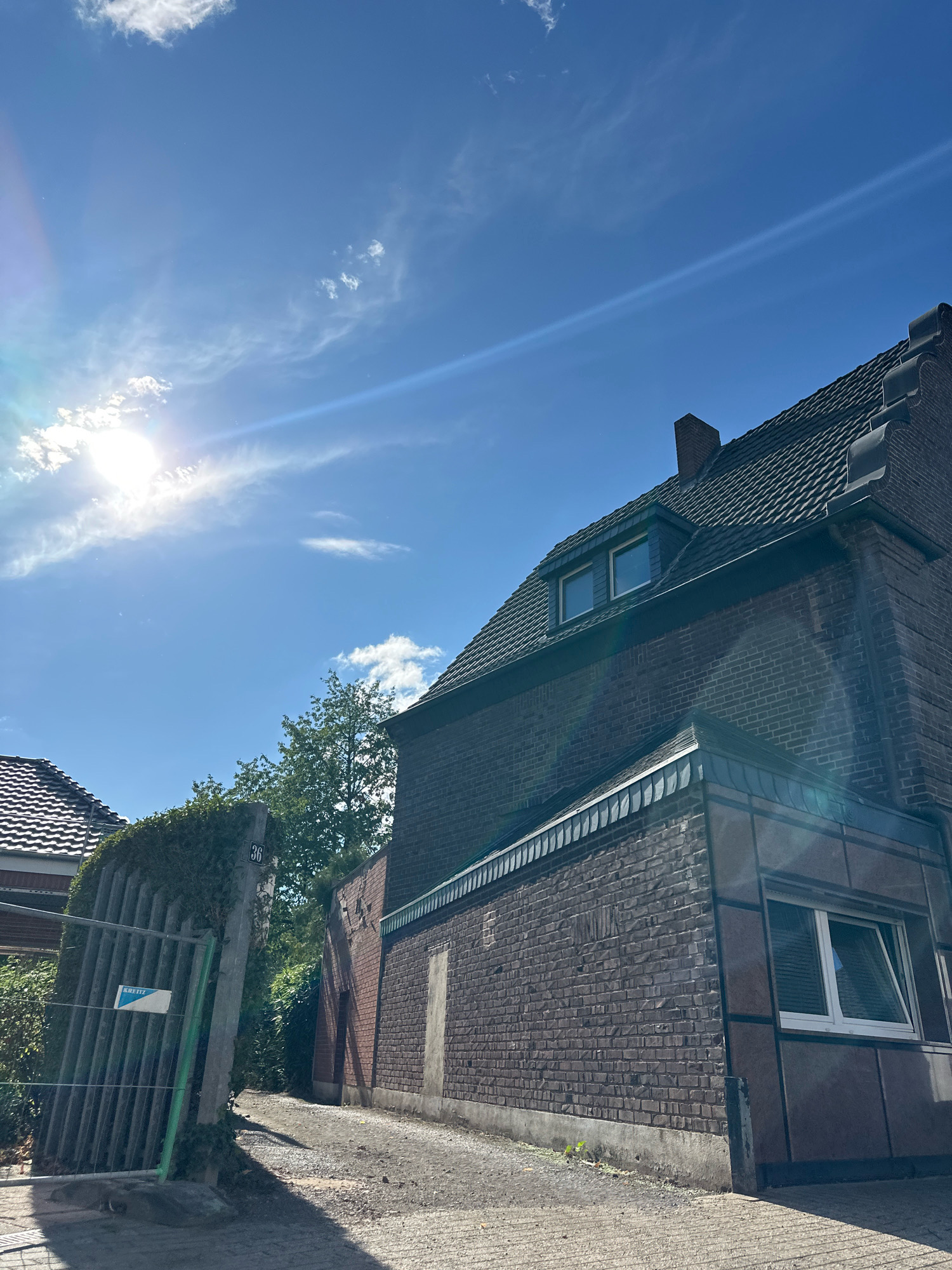 The image captures a street scene on a sunny day in Krefeld, Germany. The foreground is dominated by a paved driveway leading towards a brick building. To the left, a metal fence covered with greenery stands at an angle, with a sign that reads "KRETZ". A concrete post with the number "36" is visible behind the fence. The main building in the midground is constructed of dark brick and features a dark-tiled roof with dormer windows. Another building with a tiled roof is partially visible to the left. The background consists of a bright blue sky with scattered clouds and the sun, which is partially obscured by clouds and creates a lens flare effect. There are no people visible in the image.