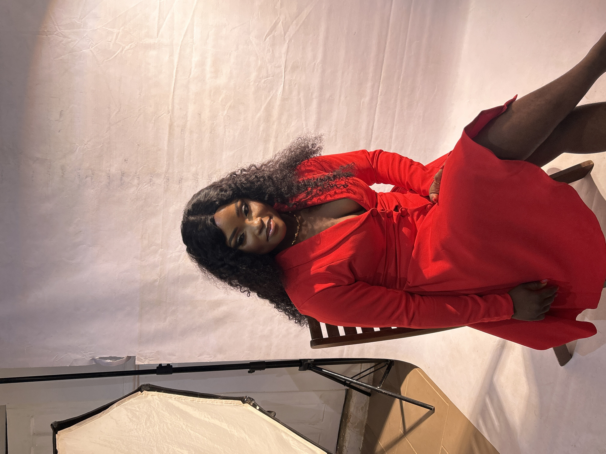 A studio shot presents a woman with dark hair, styled in loose curls, positioned reclining on a chair. She wears a bright red, long-sleeved dress, and gold jewelry is visible around her neck. Her legs are crossed.
The background consists of a white backdrop, and to the left side of the frame, there is a softbox with its stand. The location is most likely in a studio in Jos, Nigeria.