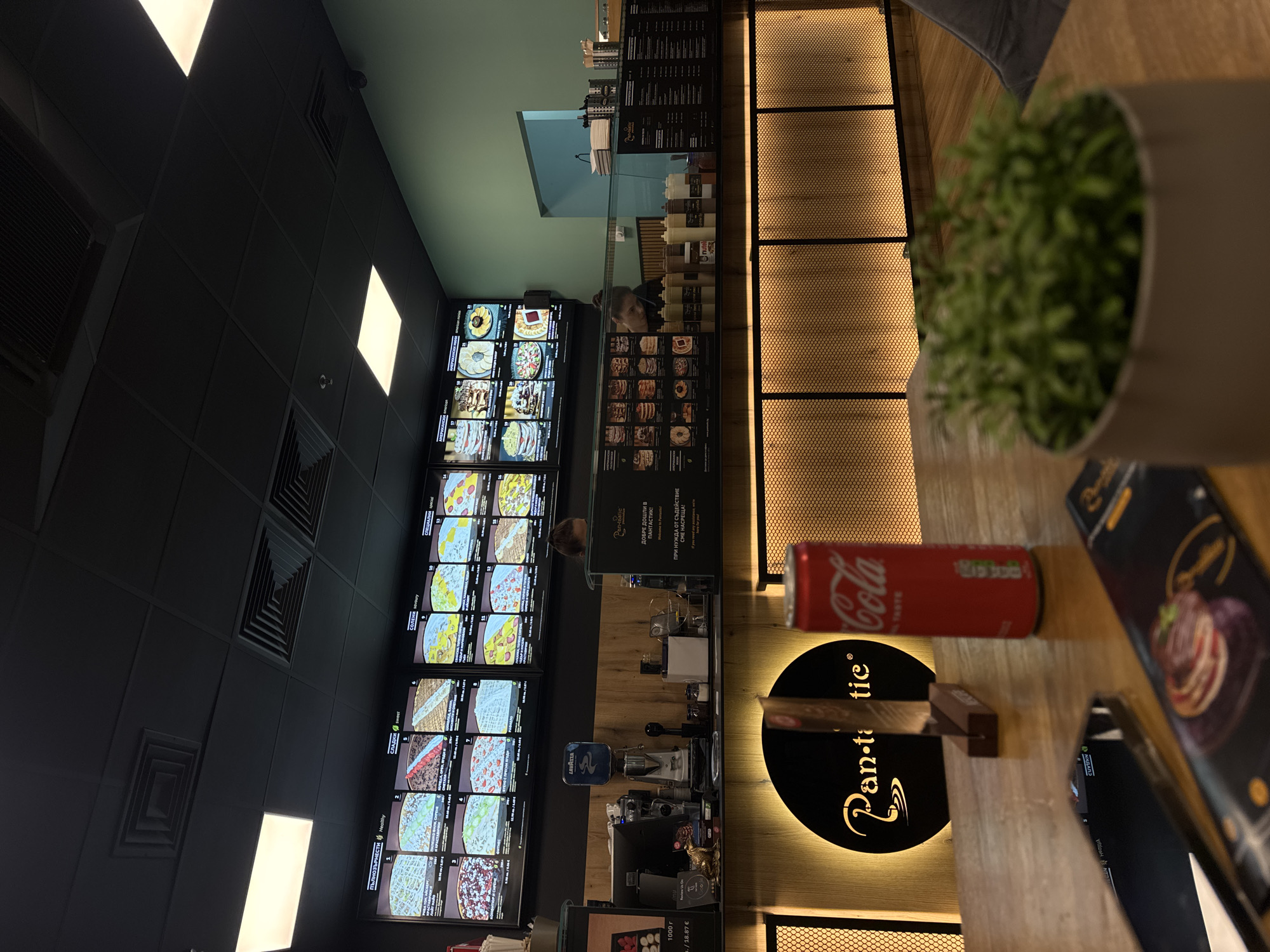The image presents an interior view of a Pan-tastic restaurant, likely located in Yambol, Bulgaria. A Coca-Cola can and promotional materials are visible on a wooden table in the foreground, partially obscured by a potted green plant. Behind the counter, the menu is displayed on multiple screens featuring images of food items. An employee is present behind the counter, which has a black and gold backlit circular logo "Pan-tastic". Various coffee machines and other food preparation equipment are also visible. The walls are dark, and the ceiling is lit with square recessed lighting. A metal mesh screen adorns part of the wall near the counter.