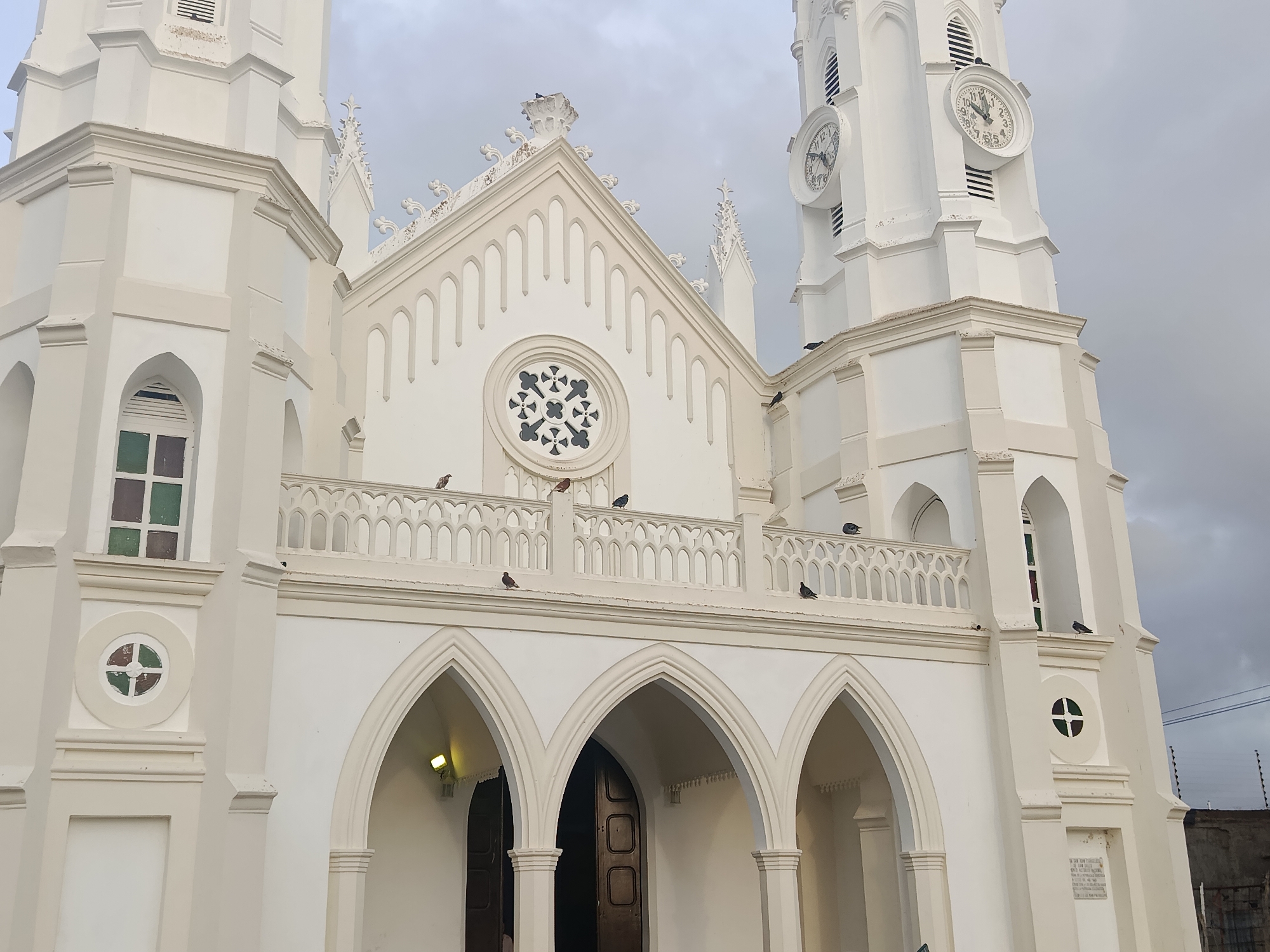 The image depicts the Iglesia de San Juan Evangelista, located in Juan Griego, Venezuela. It is a white church with a distinct Neo-Gothic architectural style. The facade features pointed arches over the entrance, a balcony with decorative railings, and tall towers with clocks. Several pigeons are perched on the balcony and other parts of the building. The sky is overcast. A fence and the side of a building can be seen to the right.