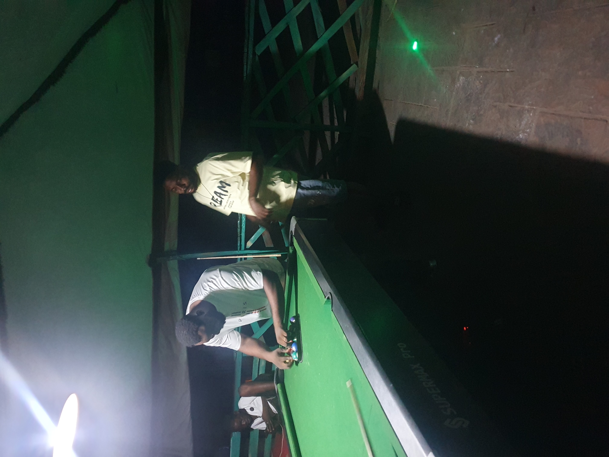 Two men are playing billiards under a covered outdoor area. One man, wearing a white t-shirt with the word "PARIS" on it, is bent over the billiard table, arranging a set of colorful balls. He has dark skin and short, dark hair. The other man, wearing a light yellow t-shirt with the word "REAM" on it and denim shorts, stands to the right of the table, looking at the balls. He also has dark skin and short, dark hair. The billiard table is green and has a black edge. The background shows a green wooden fence and a dark, shadowy environment, suggesting it is nighttime. A bright light fixture hangs from the ceiling above the players. A portion of the billiard table reads "SUPERMAX Pro". A small green light is visible on the ground to the right. The scene appears to be in Lokoja, Nigeria.