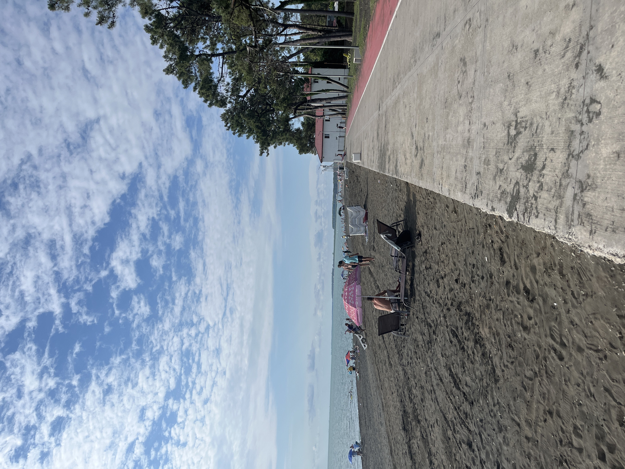 The image depicts a beach scene in Ureki, Georgia, likely during the daytime given the bright sunlight. The sky is a clear blue, dotted with scattered white clouds, suggesting fair weather. The foreground is dominated by dark, sandy beach which is quite textured with footprints and possibly small shells. Several beach chairs and umbrellas are set up on the sand, indicating a leisurely atmosphere.
In the middle ground, a paved walkway runs parallel to the beach, separated by a thin red line. This walkway appears to be a promenade or a part of a recreational area. Beyond the walkway, there are trees with lush green foliage and white buildings with red roofs, which could be hotels or residential properties. The sea is visible in the distance, appearing calm and light blue.
Several people are scattered across the beach and the walkway. Some are relaxing in beach chairs under umbrellas, while others are standing or walking. The overall mood seems relaxed and peaceful, with individuals enjoying a day at the beach. There is no visible text in the image. The orientation of the image is such that the sky is on the left and the beach and sea are on the right, suggesting the photo was taken with a vertical orientation.