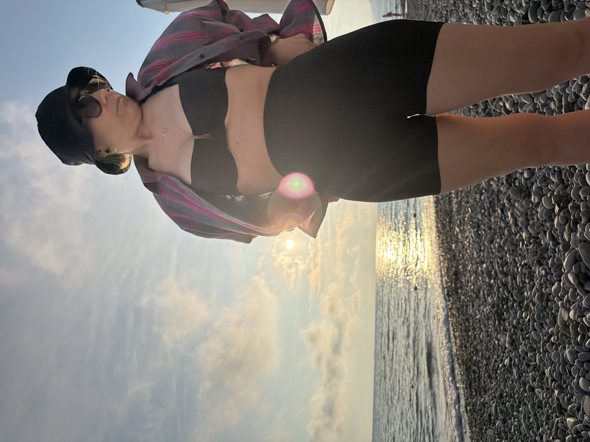 A woman stands on a pebble beach at sunset. She is wearing a black bikini top, black shorts, a plaid shirt, sunglasses, and a black bucket hat. The sun is low in the sky, casting a golden glow on the ocean and clouds. The water is calm, with small waves lapping the shore. The beach is covered in smooth, grey pebbles.