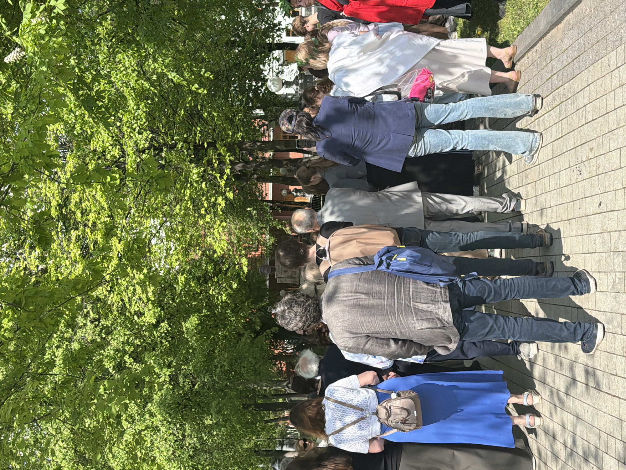 A crowd of people, mostly adults, stand on a brick sidewalk in what appears to be an outdoor urban area in Moscow, Russia. They are lined up looking towards something out of frame on the left, possibly attending or observing a ceremony or event. The clothing is varied but mostly consists of casual outerwear. Trees with dense green foliage are on the left side of the frame. The light suggests a bright, sunny day.