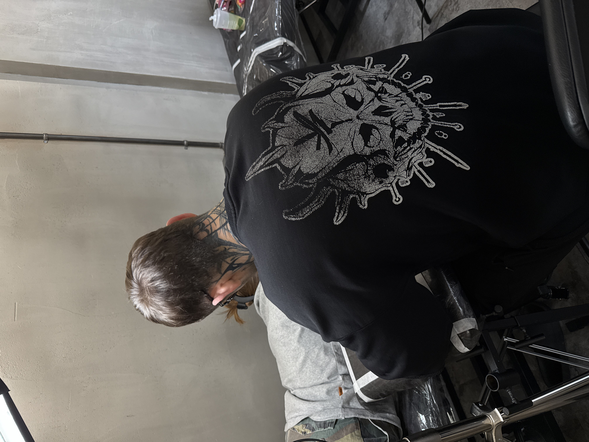 A man with short brown hair and neck tattoos is bent forward, wearing a black hoodie with a white skull design on the back, a grey shirt underneath, and camouflage pants. He is in a room with grey walls, a black metal pipe running horizontally along the wall, and a padded black chair. A wrapped object is visible near the chair. The location in Moscow, Russia, is not determinable from the image.