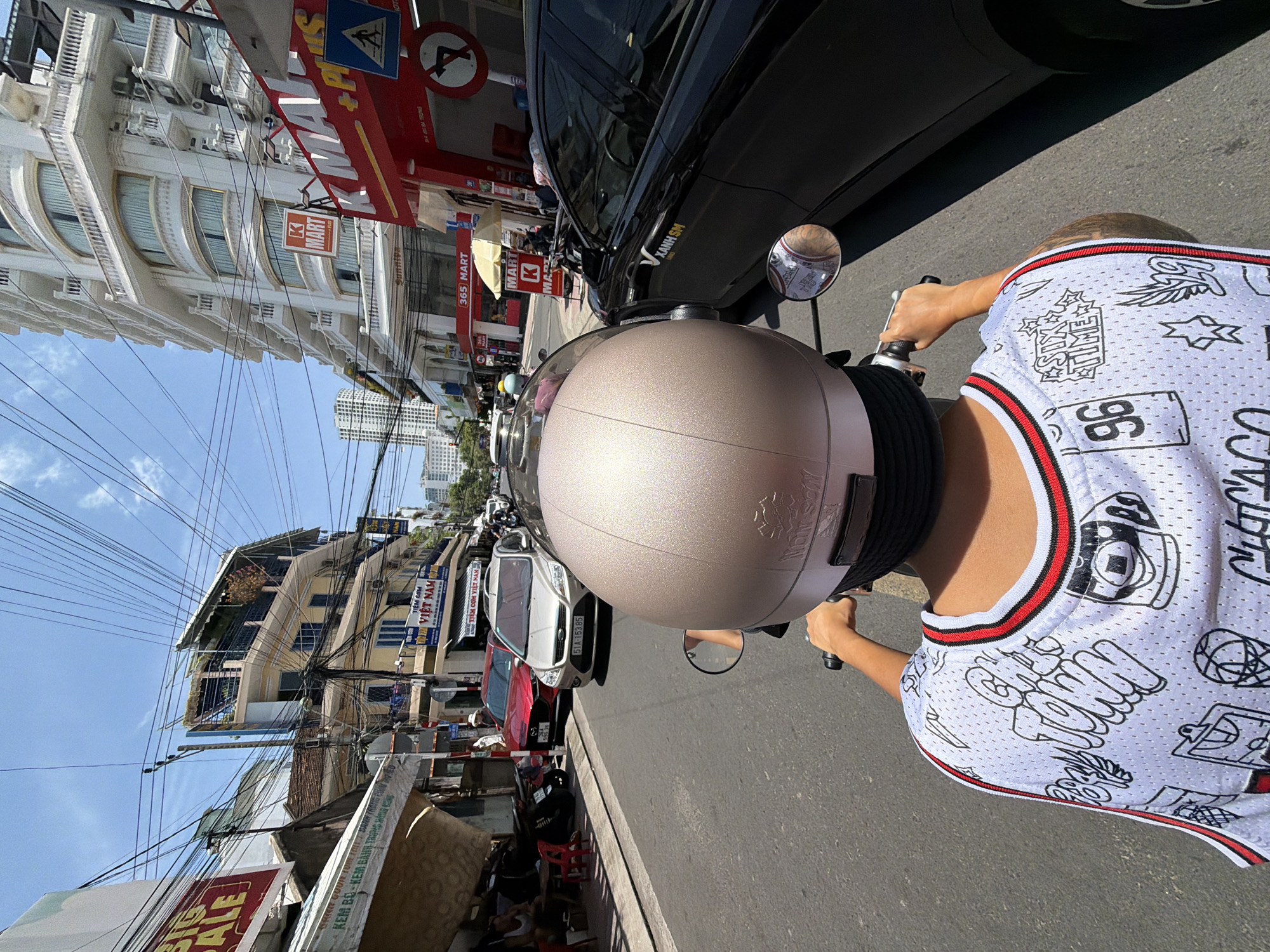 The image shows a street scene in Nha Trang, Vietnam. A person wearing a white jersey with black and red trim and a light brown helmet is riding a motorbike. The jersey has various black graphics, including the words "CITY TOWN" and the number "96". In the foreground, the person's hands are visible on the motorbike's handlebars. To the right of the motorbike, a black car is driving alongside, with "XANH SM" written on its side.
The background depicts a busy urban environment with buildings, shops, and a dense network of overhead electrical wires. On the left side of the street, there are shops with awnings and signs, including one with "BIG SALE" on it. Further down the street, a yellow building with multiple balconies is visible. On the right side, a tall white building dominates the skyline, and a red sign for "K MART" is prominent. The street is paved and appears to be a public road with other vehicles and pedestrians potentially present, although they are less visible or out of focus. The sky is clear and blue with some light clouds. The overall impression is a typical street view in a Southeast Asian city.