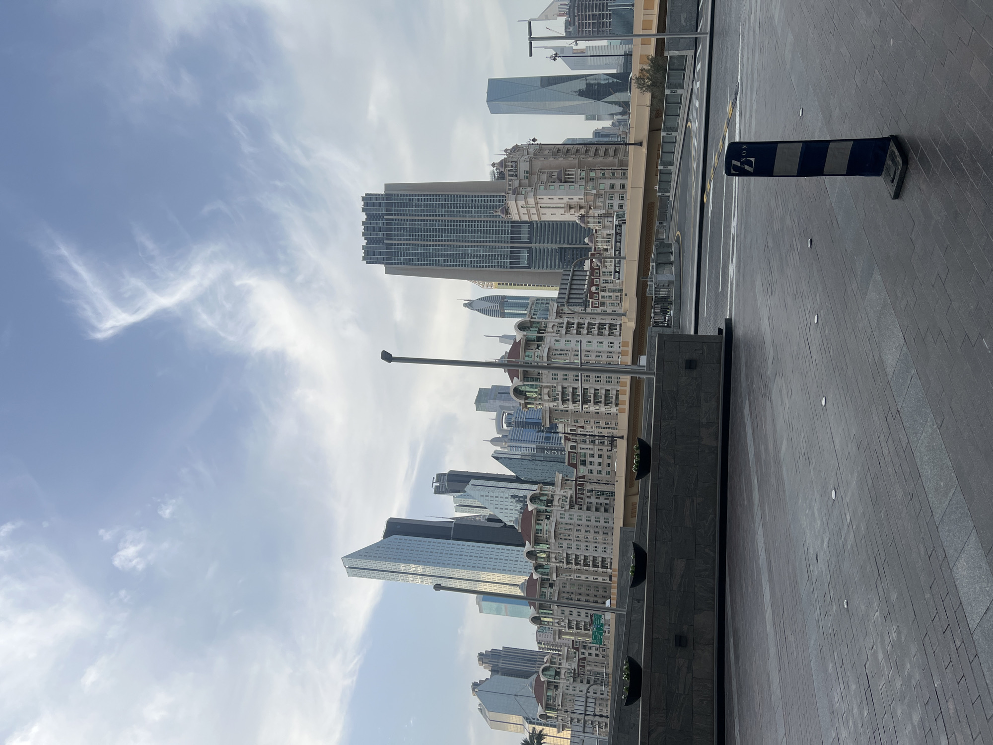 This image captures a vibrant urban cityscape under a clear daytime sky with scattered, wispy white clouds. The perspective is from ground level, looking towards a skyline of numerous buildings.
In the foreground, a wide paved area, possibly a pedestrian walkway or plaza, stretches across the bottom and right side of the frame. It features dark grey interlocking pavers and larger smooth grey tiles. Along the left edge of this paved area runs a dark grey textured wall with three black, half-moon shaped planters holding small green plants. Further along this edge, a low railing separates the walkway from an elevated section with some small trees or shrubs.
The midground and background are dominated by a diverse array of modern skyscrapers and multi-story buildings. Some prominent structures include tall, sleek glass towers reflecting the sky, as well as more traditionally-styled buildings with intricate facades, domes, and numerous windows. The buildings vary in height, creating a dynamic skyline.
Notable objects on the ground include a black and white striped bollard (traffic barrier) positioned on the right side of the paved area. On the bollard, white text is visible, which appears to be a stylized letter (resembling a Greek Xi or 'E') followed by "NON". Several streetlights with dark poles are also visible, contributing to the urban infrastructure.
The weather appears clear and pleasant, suggesting either morning or late afternoon based on the soft light. The overall scene depicts a well-developed and modern urban environment, consistent with the provided context of Dubai, United Arab Emirates. No specific activity or people are visible, presenting a calm, architectural study of the city.
