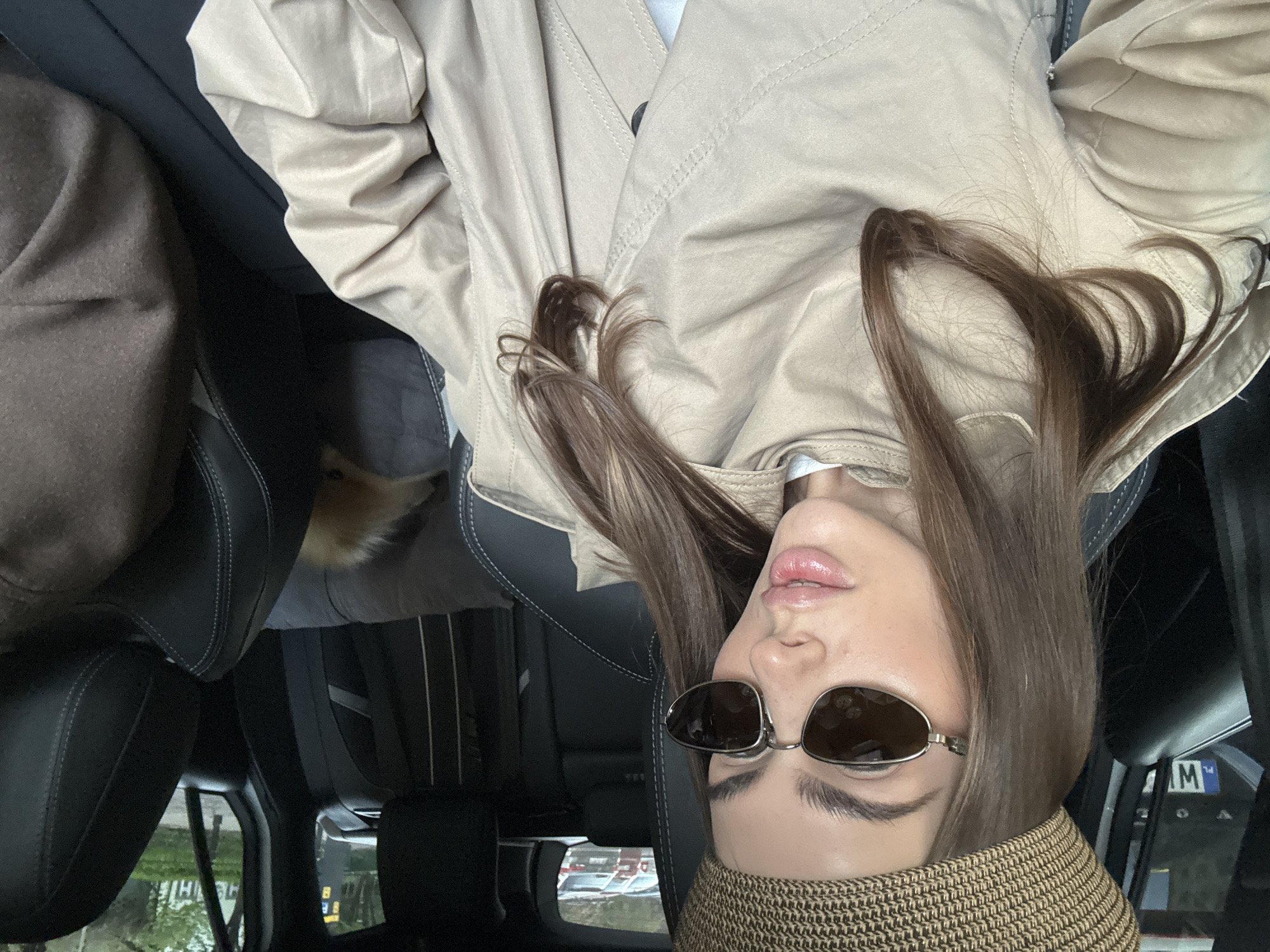 A selfie image shows a young woman with brown hair and sunglasses. She is wearing a tan hat and trench coat. The background appears to be the inside of a car. A small dog is visible in the back seat. The scene is probably in Warsaw, Poland, based on the license plate visible in the window. The car seems to be parked outside, given the street scene visible through the window.