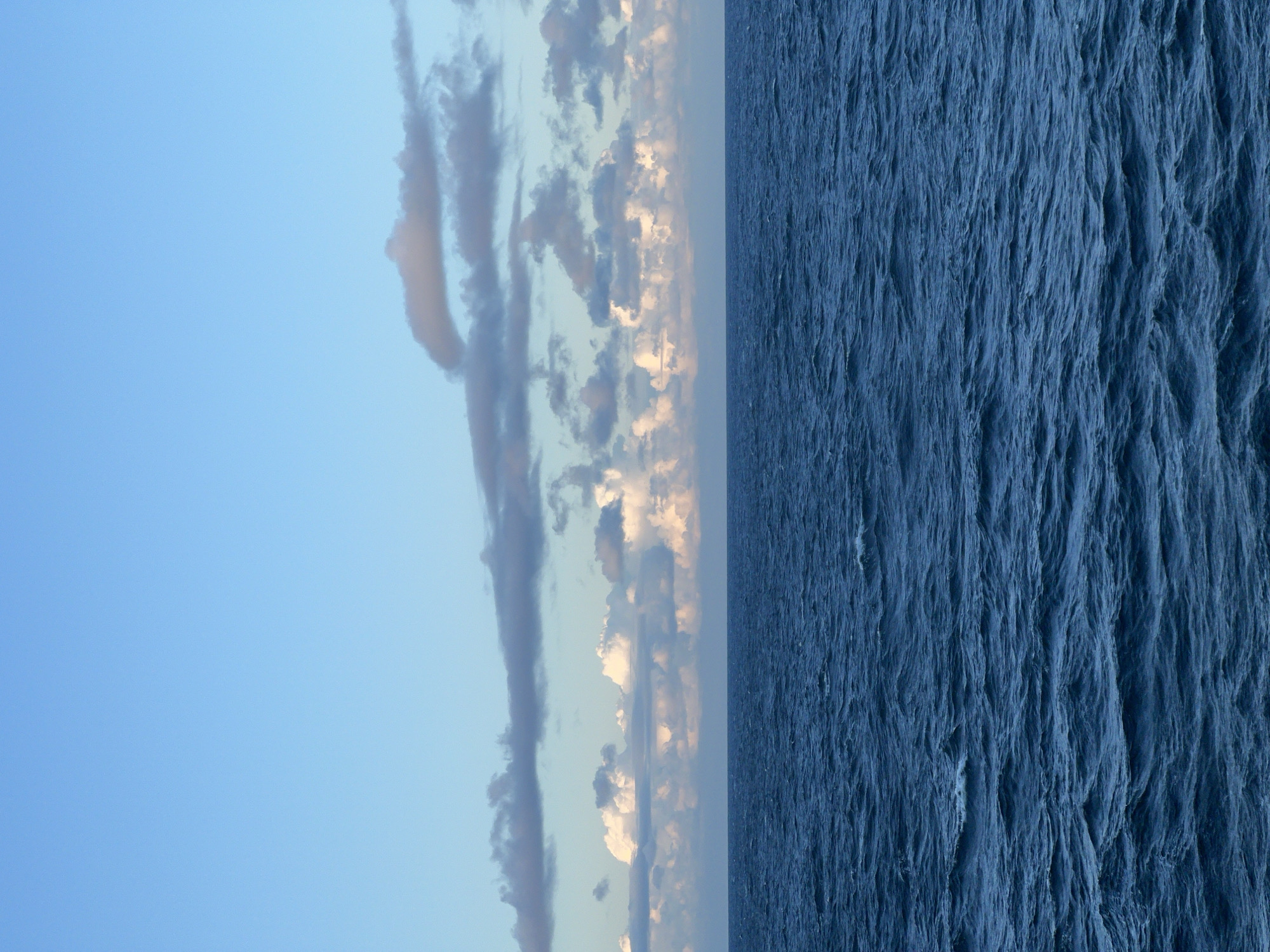 A photograph depicts the vast expanse of a dark blue ocean under a pale blue sky. The horizon line is clearly defined, separating the turbulent water from the sky. The ocean's surface is covered in numerous small waves, creating a textured and dynamic appearance. Above the water, a layer of clouds stretches across the sky. These clouds vary in density and color, with some appearing bright white and others a muted gray, suggesting a mix of sunlight and shadow. The overall scene is tranquil, with no human presence or man-made objects visible, indicating a remote or natural environment. The location is stated as Unknown, Poland, though the imagery itself does not provide specific clues to confirm or deny this.