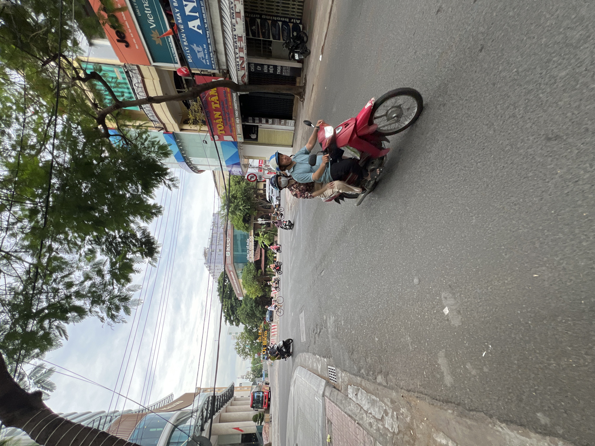 In a street scene in Nha Trang, Vietnam, two individuals are riding a red motorcycle. The person in the front, a male wearing a light blue polo shirt and a white and blue helmet, is operating the motorcycle. The passenger behind him, a female wearing a floral-patterned shirt and a white helmet, is seated. They are traveling on a paved road with a sidewalk to the left. Buildings line the right side of the street, featuring commercial signage and balconies. A large building with a rounded glass facade is visible on the far left. Other vehicles, including a red bus and several motorcycles, are present in the background, along with trees and utility poles. The sky is overcast.