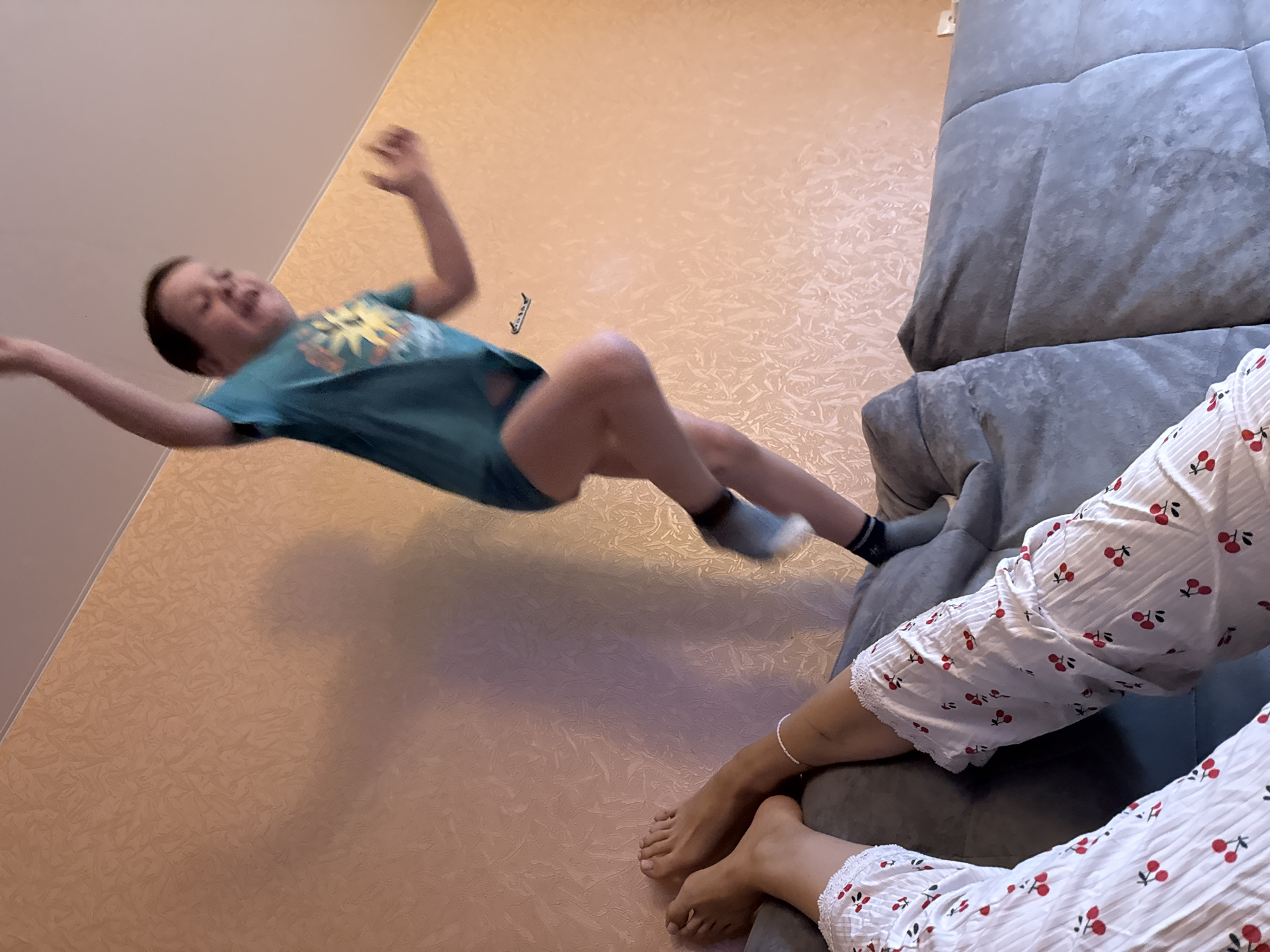 An indoor scene is captured from a low angle, showing a dynamic moment. In the center, a young child with short, dark hair is suspended in mid-air, appearing to be jumping or falling. The child is wearing a turquoise t-shirt with a yellow graphic design and dark blue shorts or underwear. Their arms are raised, and legs are bent, with white socks visible on their feet. The child's mouth is open in an expression of excitement or surprise.
To the right, a gray upholstered couch is visible, partially obscured by the frame. On the couch, the lower legs and bare feet of another person are resting. This person is wearing white pajamas adorned with a red cherry pattern and fine vertical stripes. A silver bracelet is visible on one ankle.
The background features a light orange-beige wall with a textured, abstract pattern. The top left corner shows a lighter, almost white ceiling or upper wall. A small, indistinct dark object, possibly a wall fixture, is visible on the patterned wall behind the child. The lighting suggests daytime. The overall impression is one of playful activity within a residential setting in Chaikovsky, Russia.