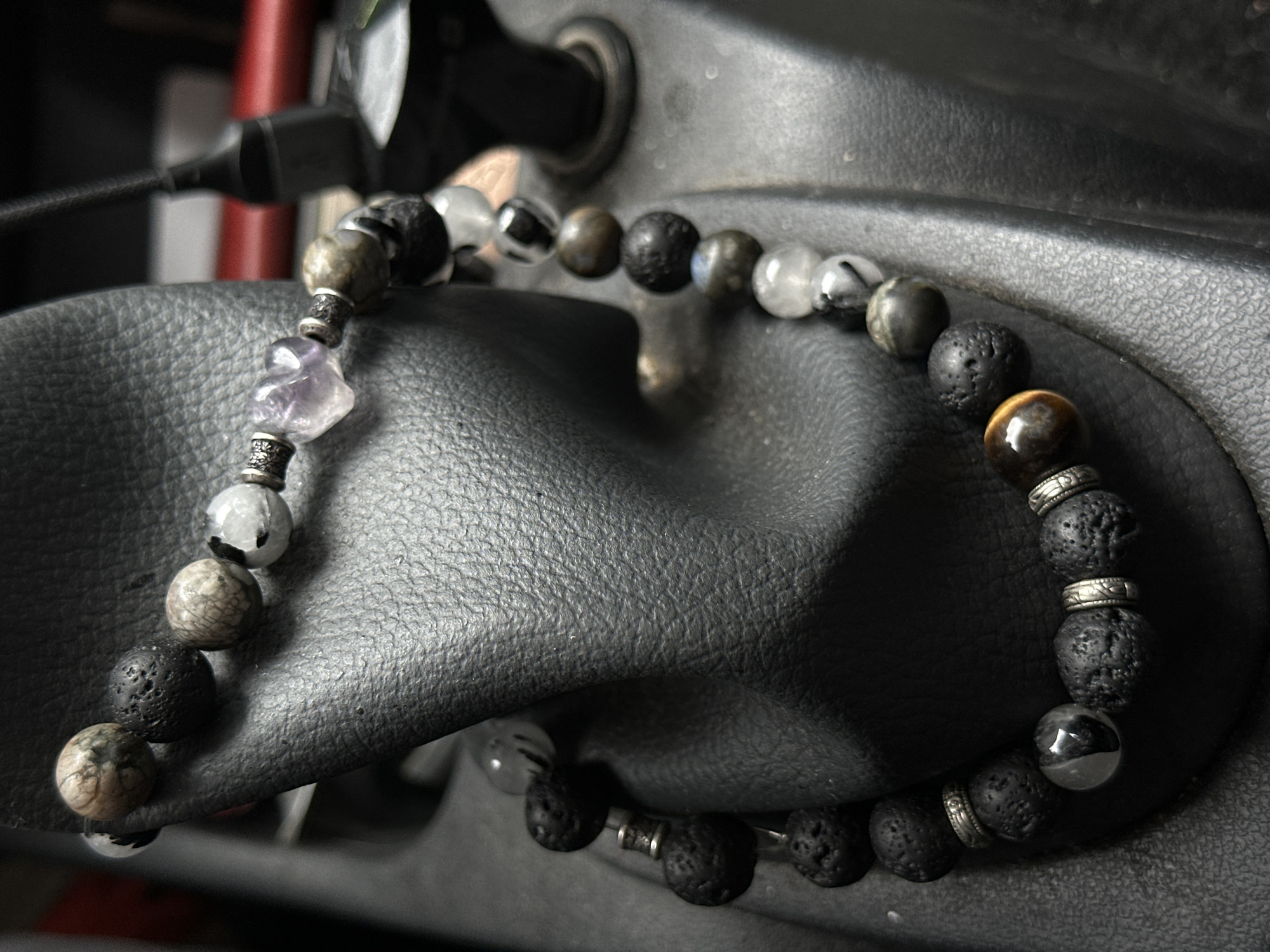 The image shows a close-up of a beaded bracelet made of black, porous lava beads, interspersed with various other beads including tiger's eye, translucent stones with dark inclusions, and faceted silver spacers. One bead at the top of the bracelet is a pale purple, heart-shaped crystal, possibly amethyst. The bracelet is draped over a black, textured surface that appears to be leather, likely part of a car's interior, such as a gear shift console. The background is out of focus but suggests an indoor environment with some electronic cables and devices visible. There are no people or animals in the image. The location is specified as Бараўлянскі сельскі Савет, Belarus, but the visual evidence within the image does not directly support or refute this location.