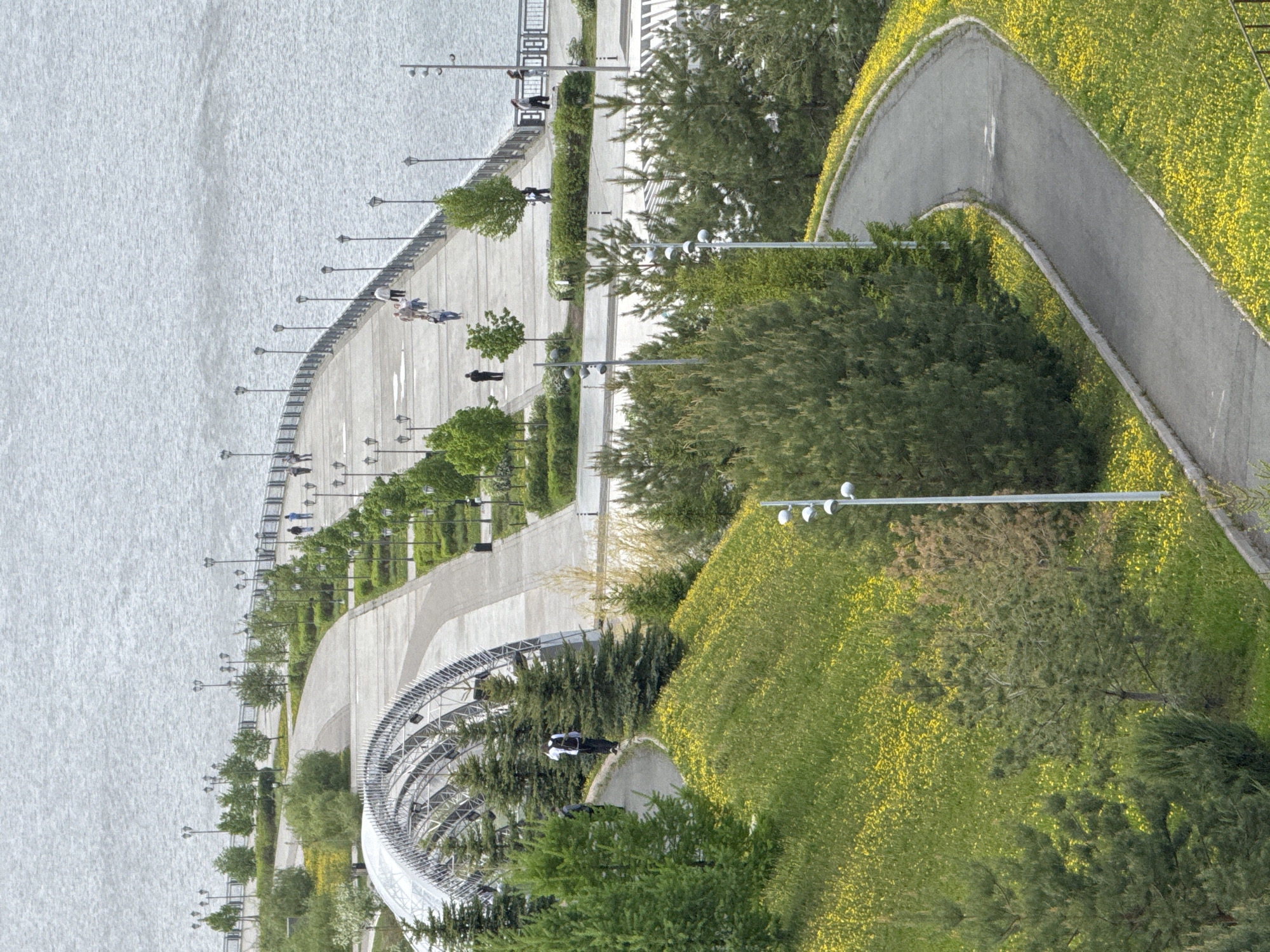 This image shows a scenic aerial view of Kazan, Russia, capturing a waterfront area with prominent walking paths and green spaces. The waterfront features a concrete promenade dotted with lamp posts and trees, where a few people are strolling. A paved path curves through grassy areas filled with yellow flowers, leading into denser tree cover. A distinctive, arched metal structure, possibly part of a park feature, sits near the pathway. In the background, there is a body of water with light, rippling waves, which is likely part of the Volga River.