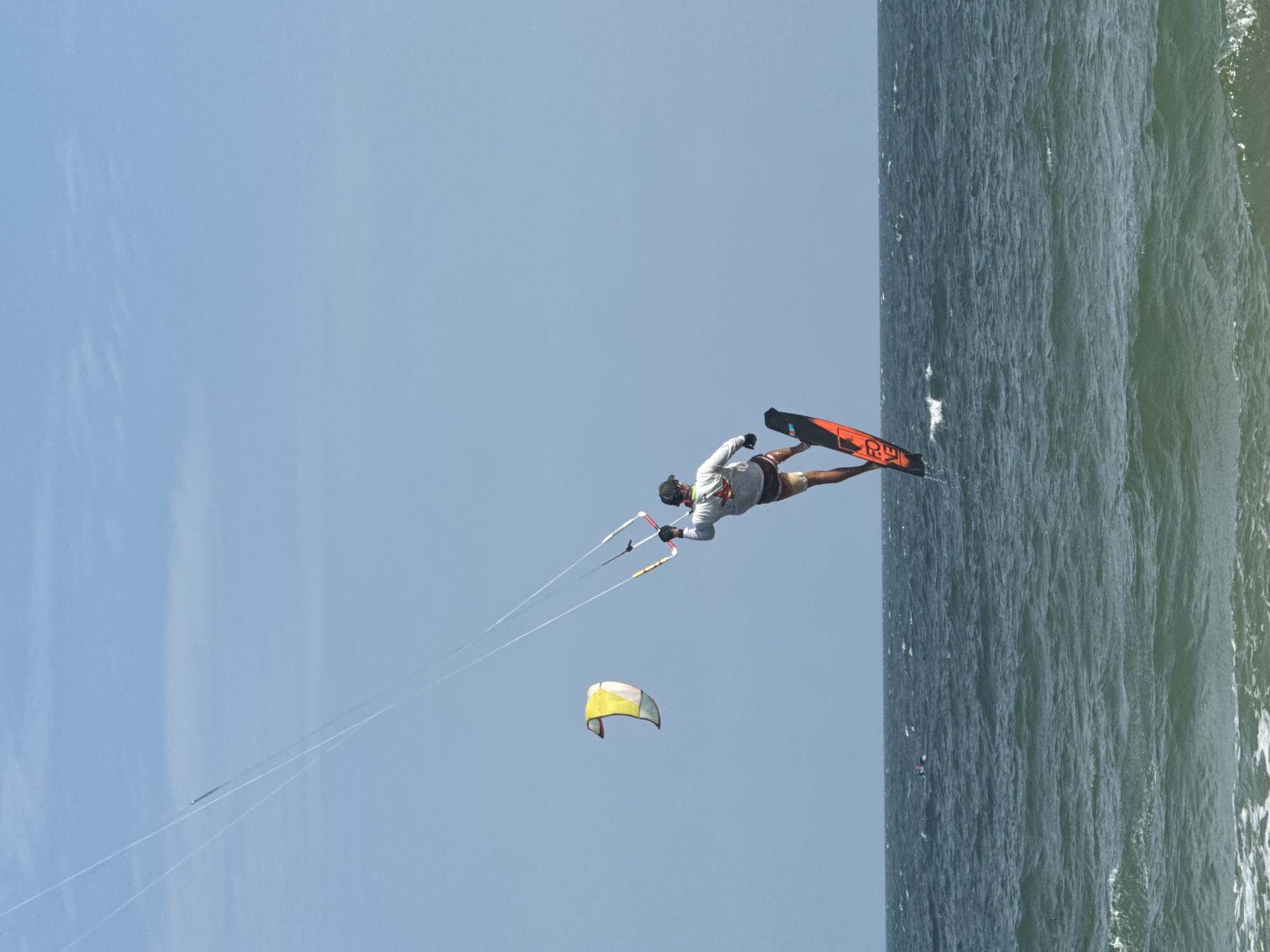 A kitesurfer is captured mid-air over the ocean in Mũi Né, Vietnam. The kitesurfer, wearing a white long-sleeved shirt and red shorts, is on a black and orange board, performing a trick. Above and to the left, another kite, yellow and white, is visible against a clear blue sky. The ocean is choppy, with waves and whitecaps. In the distance, a few other people are visible on the water, possibly also kitesurfing or windsurfing. The image depicts a dynamic scene of extreme sports in a coastal environment.