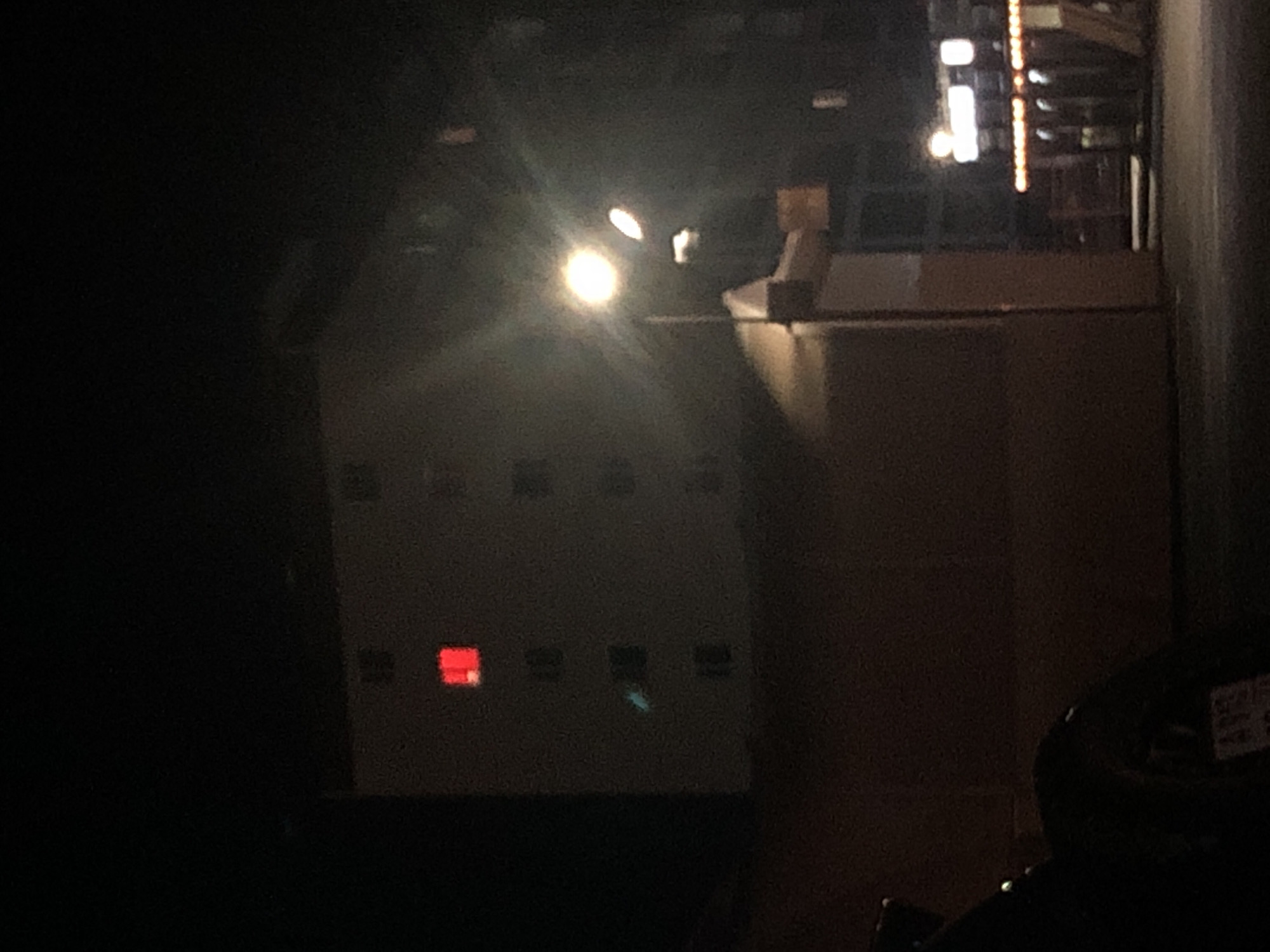 The image depicts a dimly lit interior scene. The primary subject appears to be a dark brown cabinet with a series of square indentations on its front. One of these indentations contains a bright red light. To the right of the cabinet, a lighter brown structure is visible, possibly a counter or desk. Above and behind the cabinet, a bright, blurry light source emanates, possibly a spotlight or a very bright lamp, casting lens flares. In the background, there are indications of more structured elements, suggesting a larger building or stage setup, with some vertical lines and what appears to be a series of lights or illuminated elements arranged in a vertical line. The overall atmosphere is dark, with limited visibility and a sense of being indoors, possibly in a backstage area, control room, or a space with stage lighting. There are no people clearly visible, though the presence of what looks like a stage or performance area implies human activity. The time of day is indeterminate due to the interior lighting, but the darkness suggests it is either night or the space is intentionally kept dark. There is no visible weather information. No text is discernable in the image.