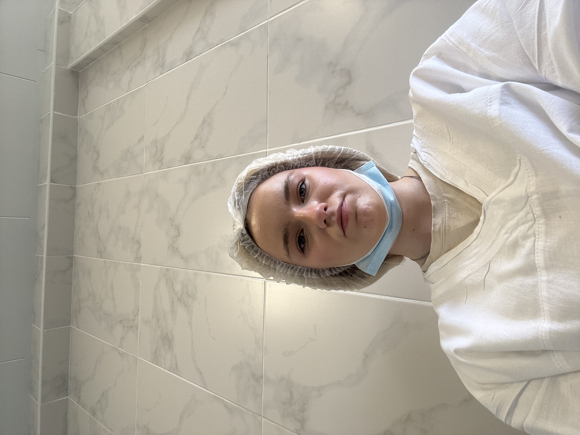 A young person, likely female, is depicted in a close-up, low-angle portrait, looking directly at the camera. She wears a disposable white bouffant cap over her hair and a light blue medical mask, which is pulled down to rest below her chin. Her attire includes a light-colored undershirt, possibly beige, topped by a loose-fitting, white long-sleeved garment, potentially a medical smock or coat. Her expression is neutral and composed.
The background features clean, light-colored tiled walls with a subtle grey marbled pattern. The rectangular tiles are uniformly arranged with thin grout lines. A corner of the room is visible on the left, indicating an indoor setting. The clinical appearance of the tiled environment, along with the subject's cap and mask, strongly suggests a professional medical or cosmetic facility, such as a doctor's office, clinic, or beauty salon. There is no visible text in the image. This scene is situated in Saint Petersburg, Russia.