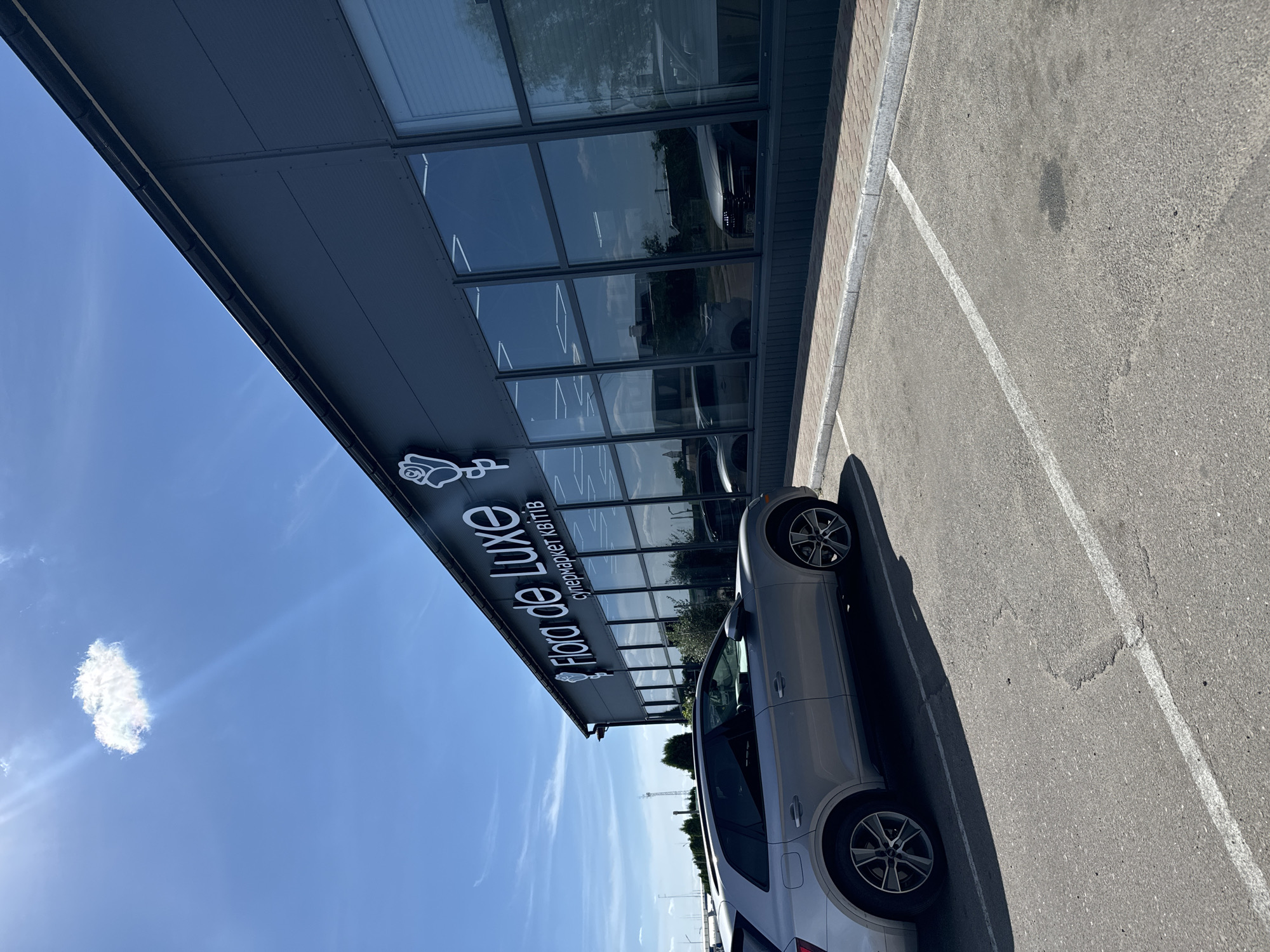 The image shows a silver car parked in a lot next to a building with large windows. The building has a sign that reads "Flora de Luxe" with an image of a flower, and beneath it, in smaller text, "супермаркет квітів". The lot is paved with asphalt and has white parking lines. The sky is blue with a few white clouds, and the sun is shining, suggesting it is daytime. The car's shadow is cast on the ground, indicating the sun is to the left of the frame. The scene appears to be a commercial area, possibly a flower shop or market. The overall mood is bright and sunny.