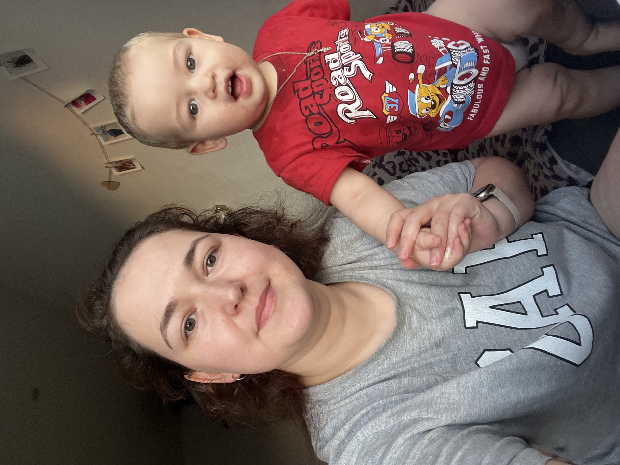 The image shows a close-up selfie of a woman and a young child, likely taken indoors. The woman is on the left, wearing a gray t-shirt with a large white "GAP" logo. She has brown curly hair and is holding the child's hand. The child is on the right, wearing a red t-shirt with cartoon characters and the text "Road Sports". The child has blonde hair and an open mouth, looking towards the camera. In the background, there is a wall with a string of small photographs clipped to it. The lighting suggests it is daytime, with sunlight casting shadows on the wall. The overall scene appears to be a casual, personal moment. There is no information in the image to indicate the location is in Chelyabinsk, Russia.