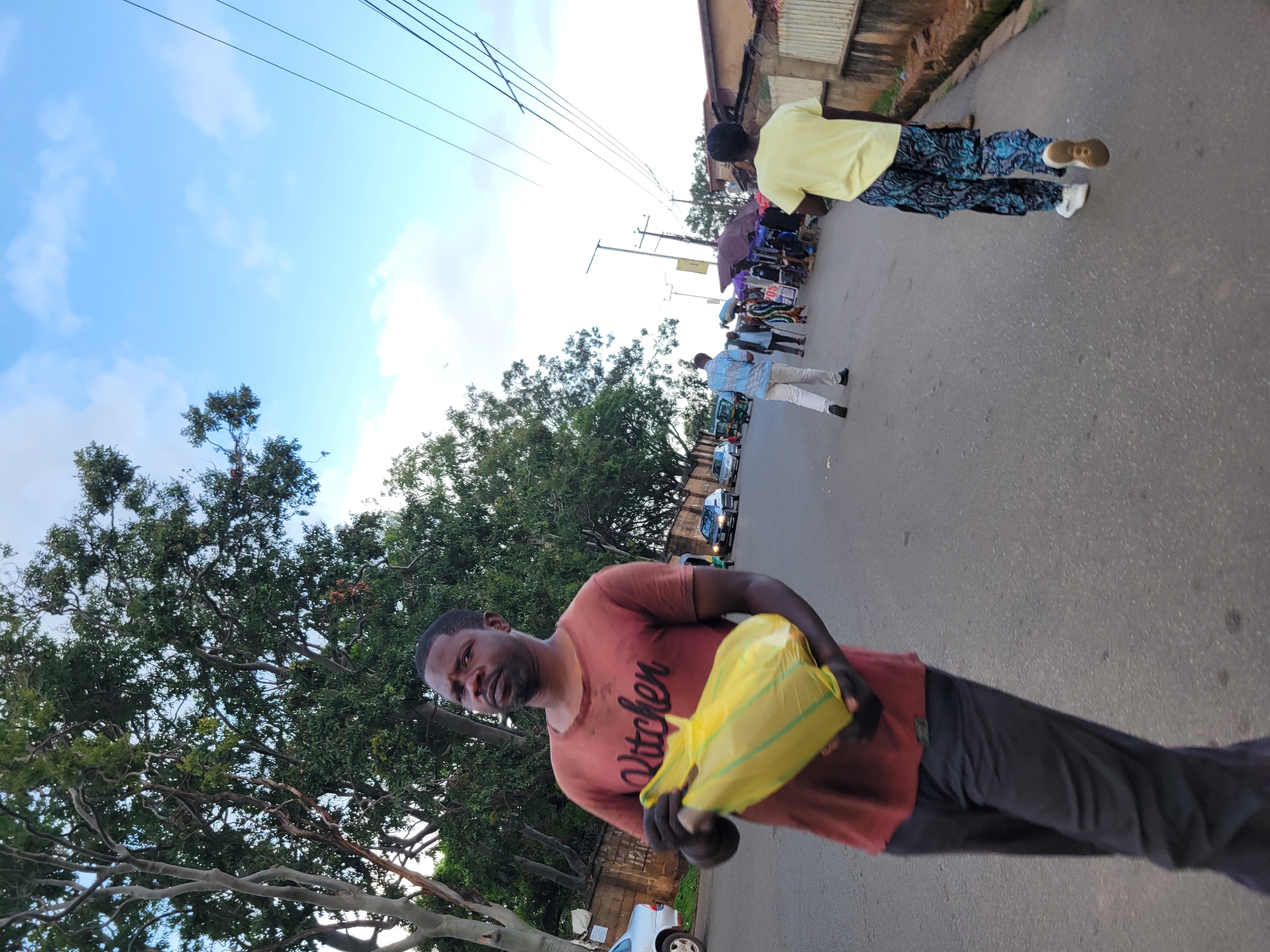 The image captures a street scene in Jos, Nigeria, with a focus on a man in the foreground carrying a bright yellow plastic bag. He is wearing a rust-colored t-shirt with the word "Kitchen" printed on it in black lettering. He has a beard and appears to be looking directly at the camera with a neutral expression. He is holding the yellow bag with both hands, suggesting he might be carrying groceries or purchased items.
In the background, other people are visible walking along the paved street, some of whom appear to be near market stalls or shops. One person in the mid-ground is wearing a light yellow shirt and patterned pants. Several umbrellas are open, possibly providing shade from the sun. Vehicles, including what looks like a small bus or van, are also present on the road. Trees line the side of the street, and a brick wall is visible in the background, along with some corrugated metal structures.
The sky is blue with scattered white clouds, indicating it is likely daytime, perhaps late morning or afternoon. The overall lighting suggests fair weather. The image is oriented vertically, with the subjects appearing sideways.