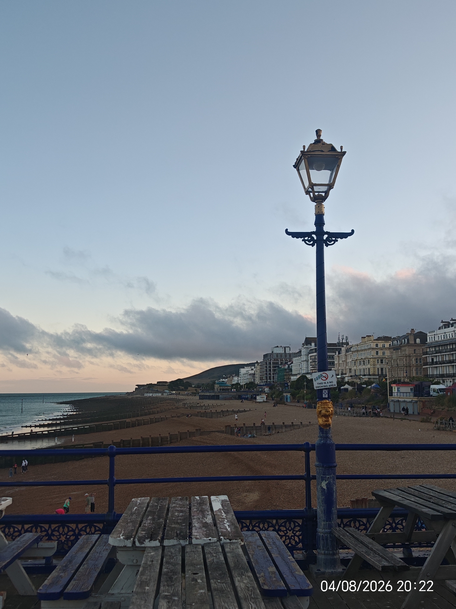 This image depicts a view of Eastbourne beach and promenade in Eastbourne, United Kingdom. In the foreground, there are wooden picnic tables and benches, with a blue railing behind them. A tall, ornate blue lamppost with a golden lion's head at its base stands prominently. A "NO SMOKING AREA" sign is attached to the lamppost. Beyond the railing and tables, the pebble beach stretches towards the sea, with wooden groynes marking the shoreline. The water is calm, and the sky is a pale blue with scattered clouds, tinged with pink at the horizon, suggesting it is either sunrise or sunset. Buildings line the promenade in the background, forming a coastal townscape. Several people are visible on the beach and promenade, engaged in various activities. The timestamp in the lower right corner indicates the image was taken on August 4, 2026, at 20:22.