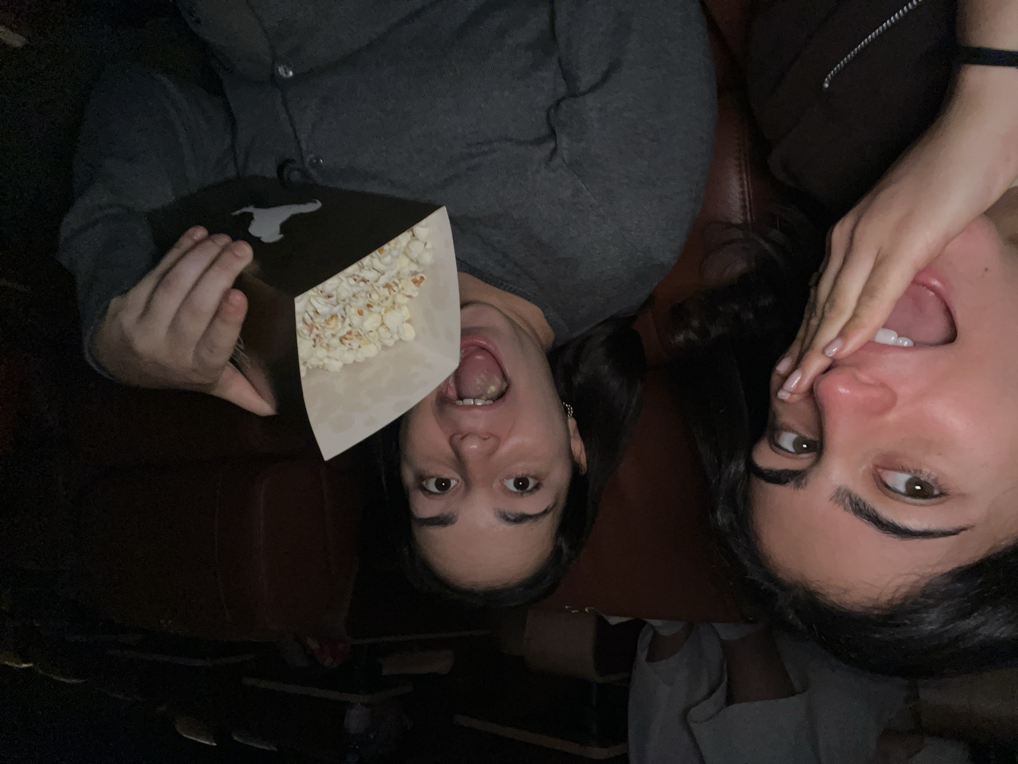 Two women are in a dark setting, possibly a movie theater. One woman is holding a box of popcorn, with her mouth open. The second woman is lying next to her. The location is unknown and there is no evidence that it is in Paris, France.