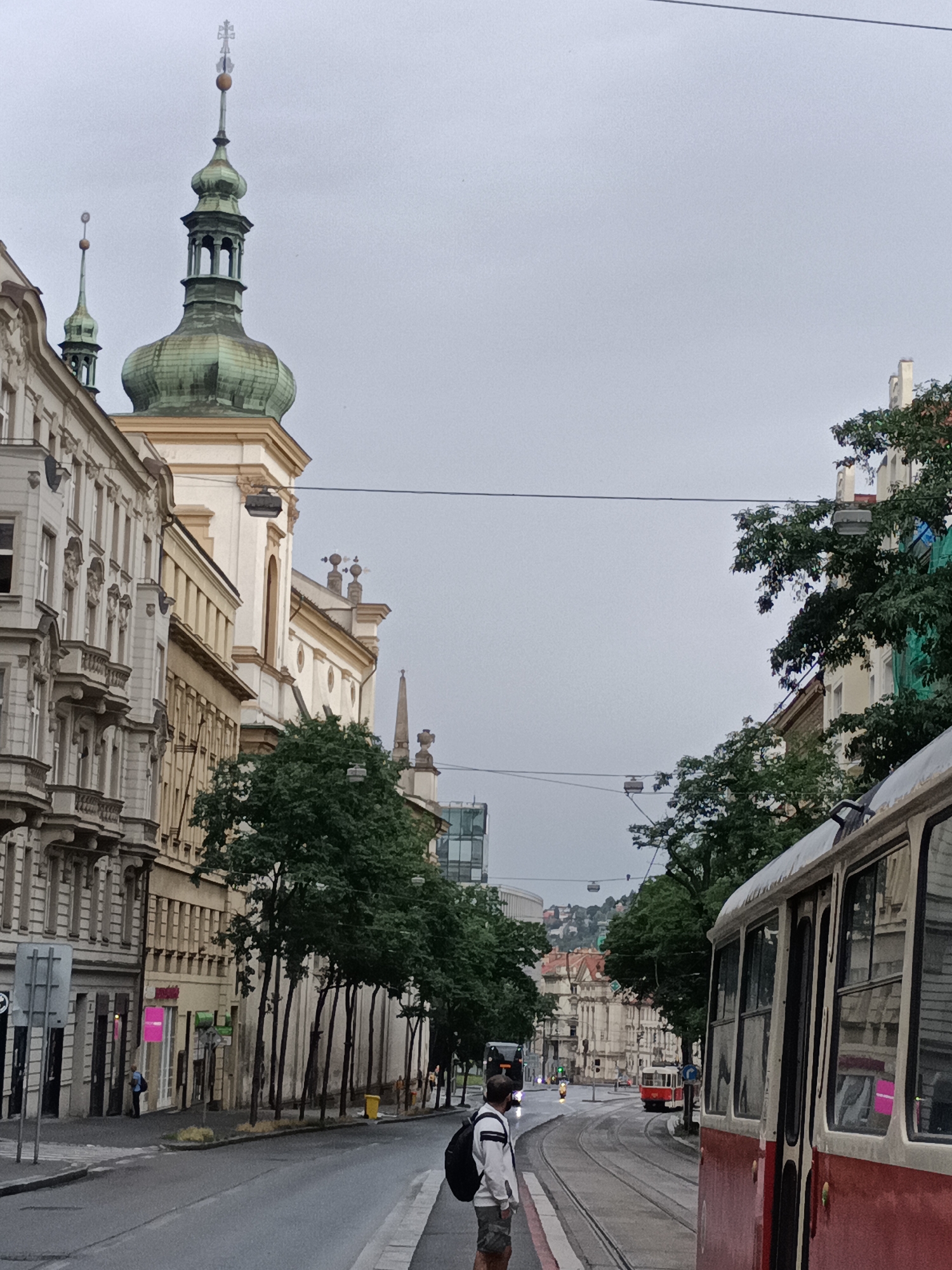 The image shows a street scene in Prague, Czechia, likely near the Strahov Monastery. A tram is prominent in the foreground on the right, with its red and white exterior visible. In the background, a man stands facing away from the camera, looking down the street. Other trams and a bus are visible further down the road. On the left side of the street are several buildings with ornate facades and balconies, characteristic of Prague's architecture. A church spire or tower with a green dome is visible above the buildings. Trees line the street, and the sky is overcast. The street appears to be cobblestoned or paved, with tram tracks running down the center.