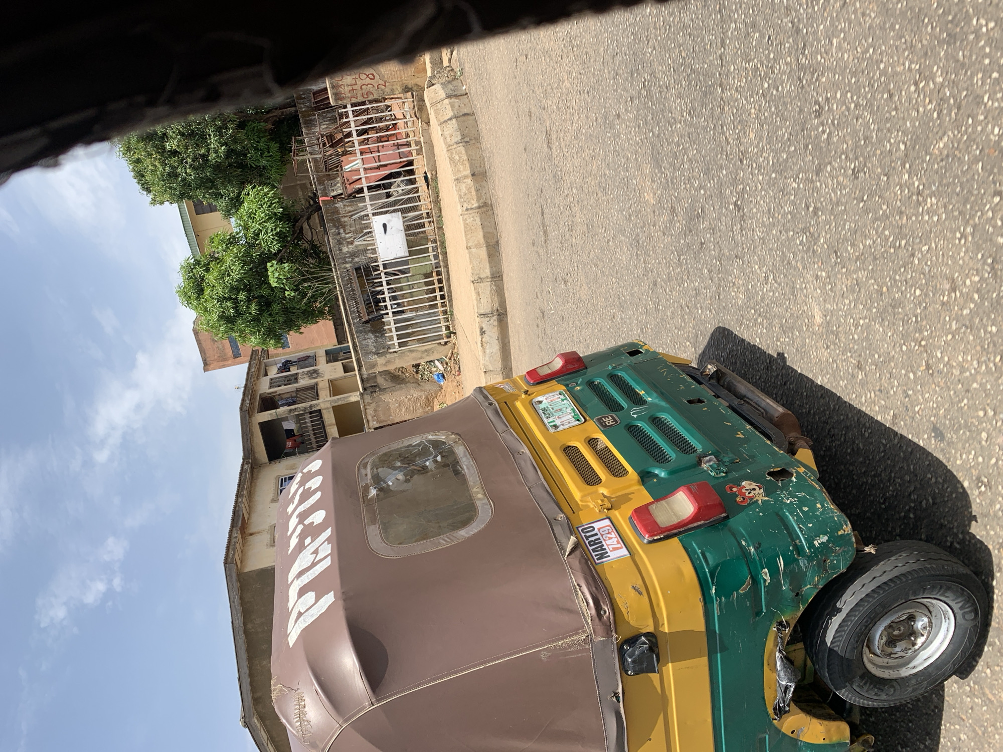 The image shows a three-wheeled vehicle, a "keke," in Rafin Par, Nigeria. The keke is green and yellow with a brown canvas roof. It is parked on an asphalt road beside a curb. The rear of the keke is visible, showing a license plate and a NARTO sticker. To the left are buildings of tan concrete and a metal fence. A tree with green foliage is present. The sky is visible in the upper-left and is pale blue with some cloud cover. The image is taken from inside another vehicle. The overall condition of the keke appears worn and used.