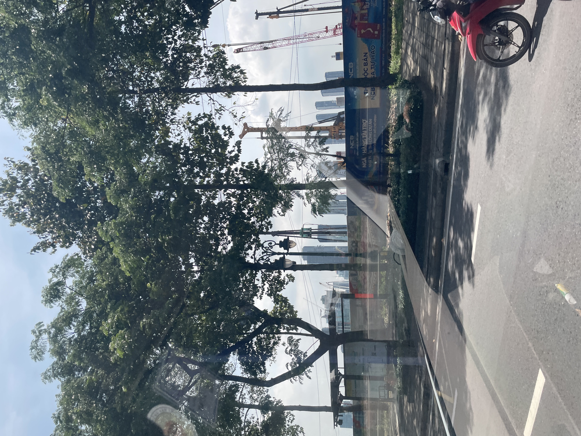 The image shows a street view in Thủ Đức City, Vietnam, with a reflection of trees and the sky on the windshield of a vehicle. In the foreground, a red motorcycle is parked on the right side of the road. The street has white lane markings. Beyond the street, there is a sidewalk lined with trees. In the background, several tall buildings and construction cranes are visible, indicating ongoing development in the city. A billboard with Vietnamese text and logos is also present. The sky is partly cloudy.