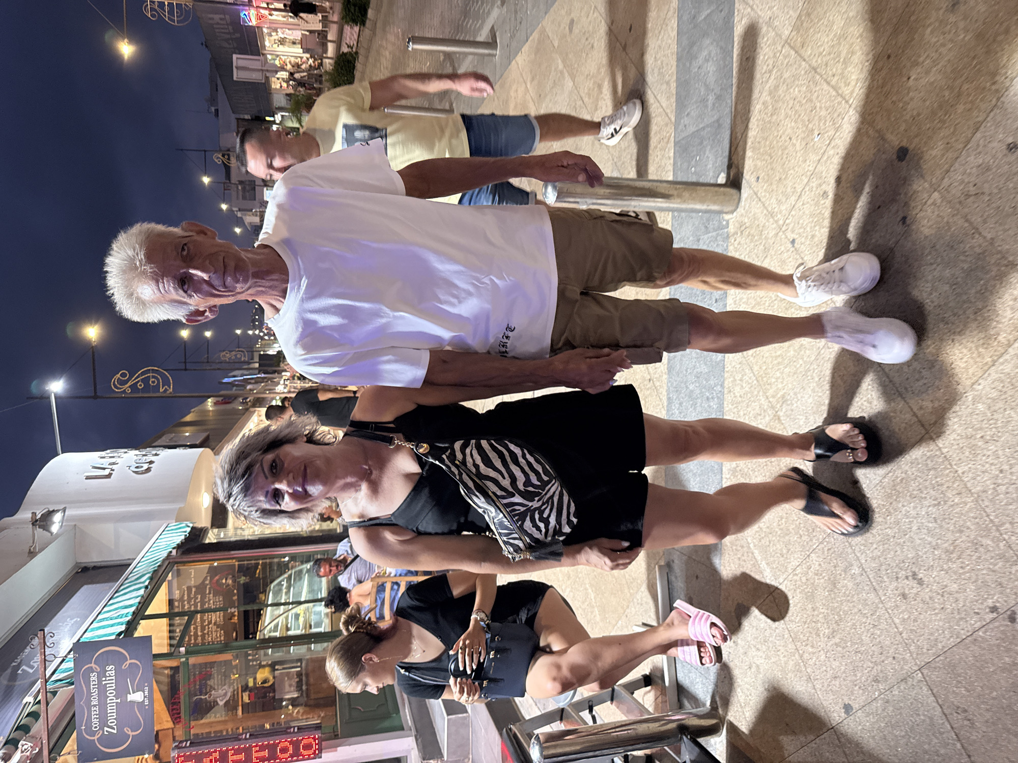The image shows a street scene at night in Ayia Napa, Cyprus. A couple, a man and a woman, are standing in the foreground, holding hands. The man is wearing a white t-shirt and khaki shorts. The woman is wearing a black dress and a zebra-print fanny pack. Behind them, a woman with blonde hair is sitting on a stool, holding a black purse. In the background, there are shops and streetlights. The overall atmosphere is casual and lively.