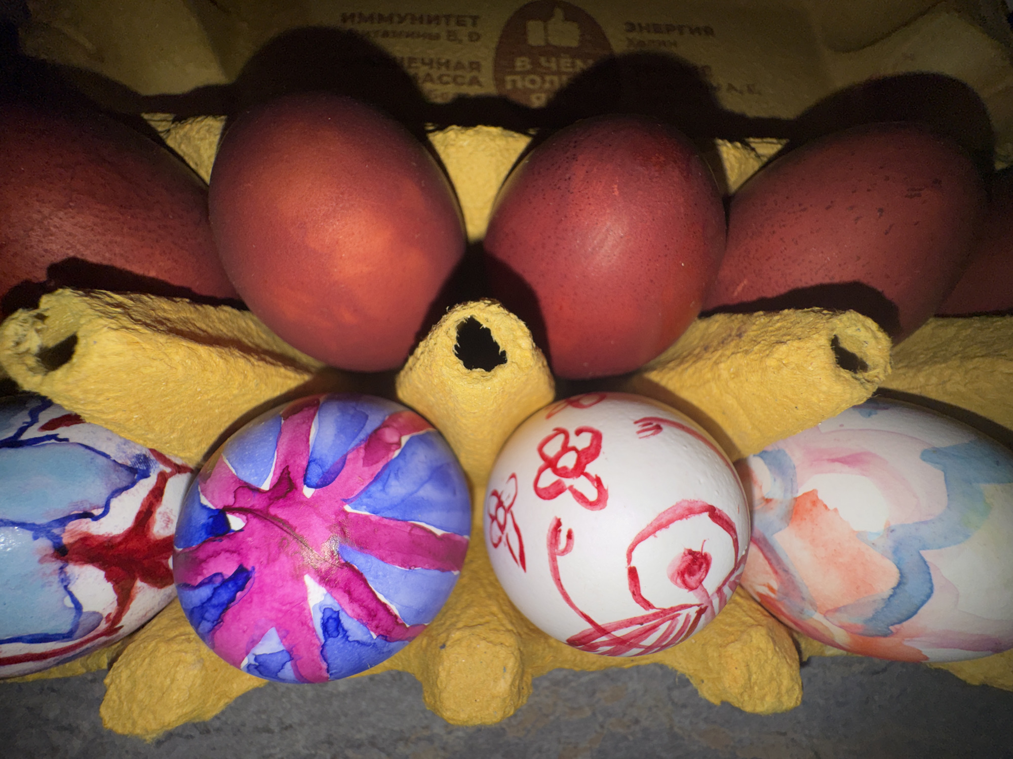 The image shows a yellow egg carton containing several Easter eggs. The top row of eggs is dyed a deep red color. The bottom row contains eggs that have been decorated with watercolor paint. The paint colors include blue, pink, red, and orange. There is writing in Russian visible on the carton. This suggests the image may have been taken in a region where Russian is spoken, such as Moscow, Russia.