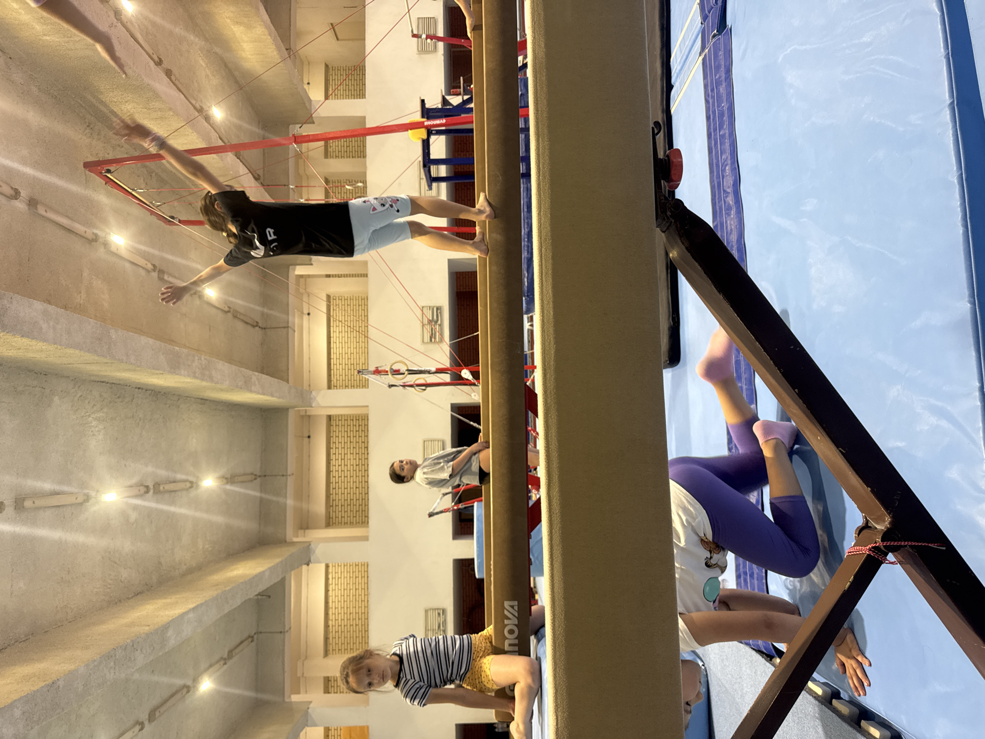 This image depicts a gymnastics training session within an indoor facility. Four young girls are visible, engaged in various activities. In the foreground, one girl with purple leggings and pink socks is on her hands and knees on a blue mat, peeking under a balance beam. To her left, another girl in a striped t-shirt and yellow shorts is kneeling on a balance beam, looking towards the camera. Further back and to the right, a third girl in a grey t-shirt and black shorts stands on a different balance beam, looking away. The tallest girl, wearing a black t-shirt with a white logo and light blue shorts with cartoon cat prints, is standing with her arms raised on a balance beam. The environment includes overhead lighting fixtures on a concrete ceiling, brick walls in the background, and gymnastics equipment such as balance beams and red apparatus frames. The overall scene suggests a dynamic and active training session. The image's metadata indicates it was taken in Sofia, Bulgaria.