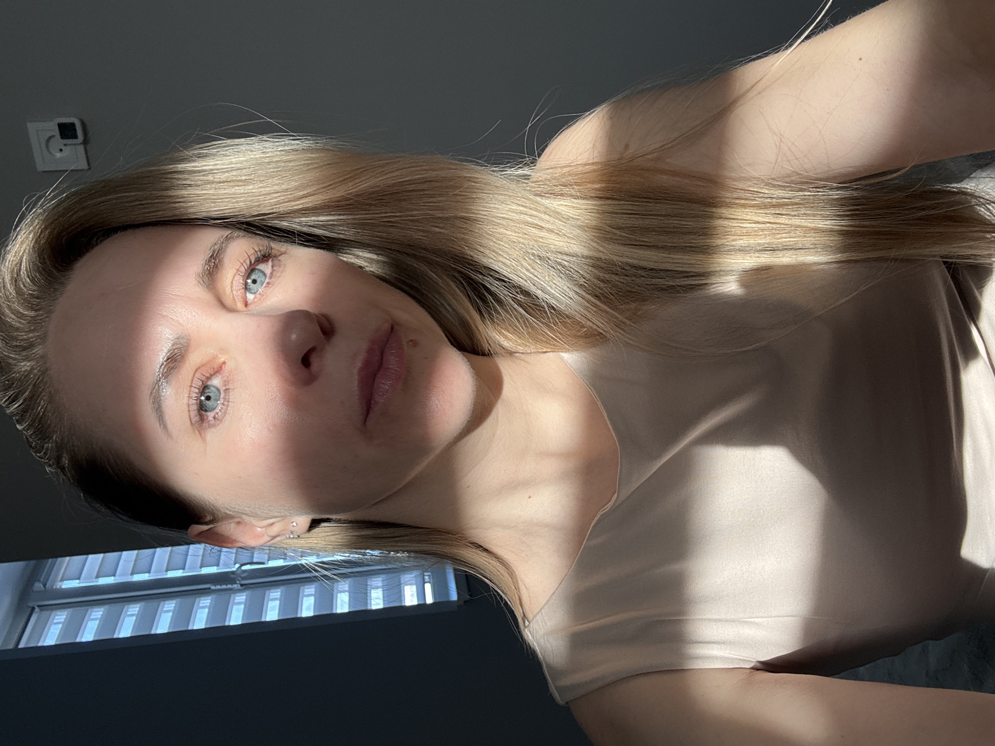 The image is a close-up, rotated selfie of a young woman indoors in Chaikovsky, Russia. She has long, straight blonde hair that cascades over her shoulder and chest, illuminated by strong, direct sunlight creating pronounced highlights and shadows. Her blue eyes are prominent, framed by visible eyelashes. Her fair complexion is partly shadowed, and a small mole is visible above her upper lip. Her left ear features several small stud earrings. She is wearing a plain, light-colored t-shirt or tank top, possibly beige or light pink.
The activity depicted is the woman taking a selfie, looking directly at the camera with a neutral, contemplative expression.
Notable details include the time of day, which appears to be daytime, likely morning or late afternoon, due to the intense, directional sunlight streaming into the room. The weather is sunny and clear, as evidenced by the sharp shadows. The setting is an indoor room, with a dark gray wall visible behind her. A white electrical wall socket, possibly with a black switch or component, is mounted on the wall above her head. In the lower left corner, a window with horizontal blinds is partially visible, indicating the source of the strong natural light. There is no visible text in the image.