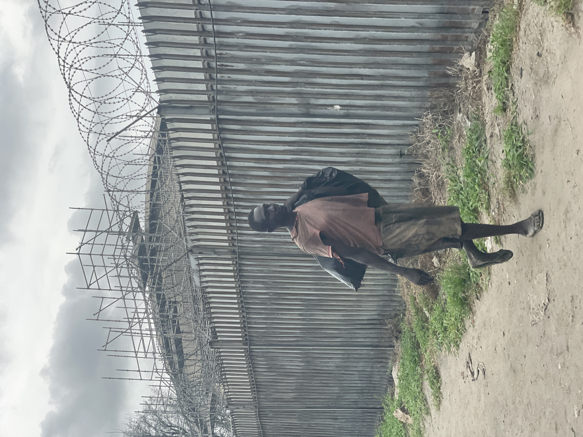 An unpaved area beside a corrugated metal wall topped with barbed wire. A man carrying a dark-colored sack walks barefoot along the unpaved area. Scaffolding supports the wall. This image was taken in Lagos, Nigeria.