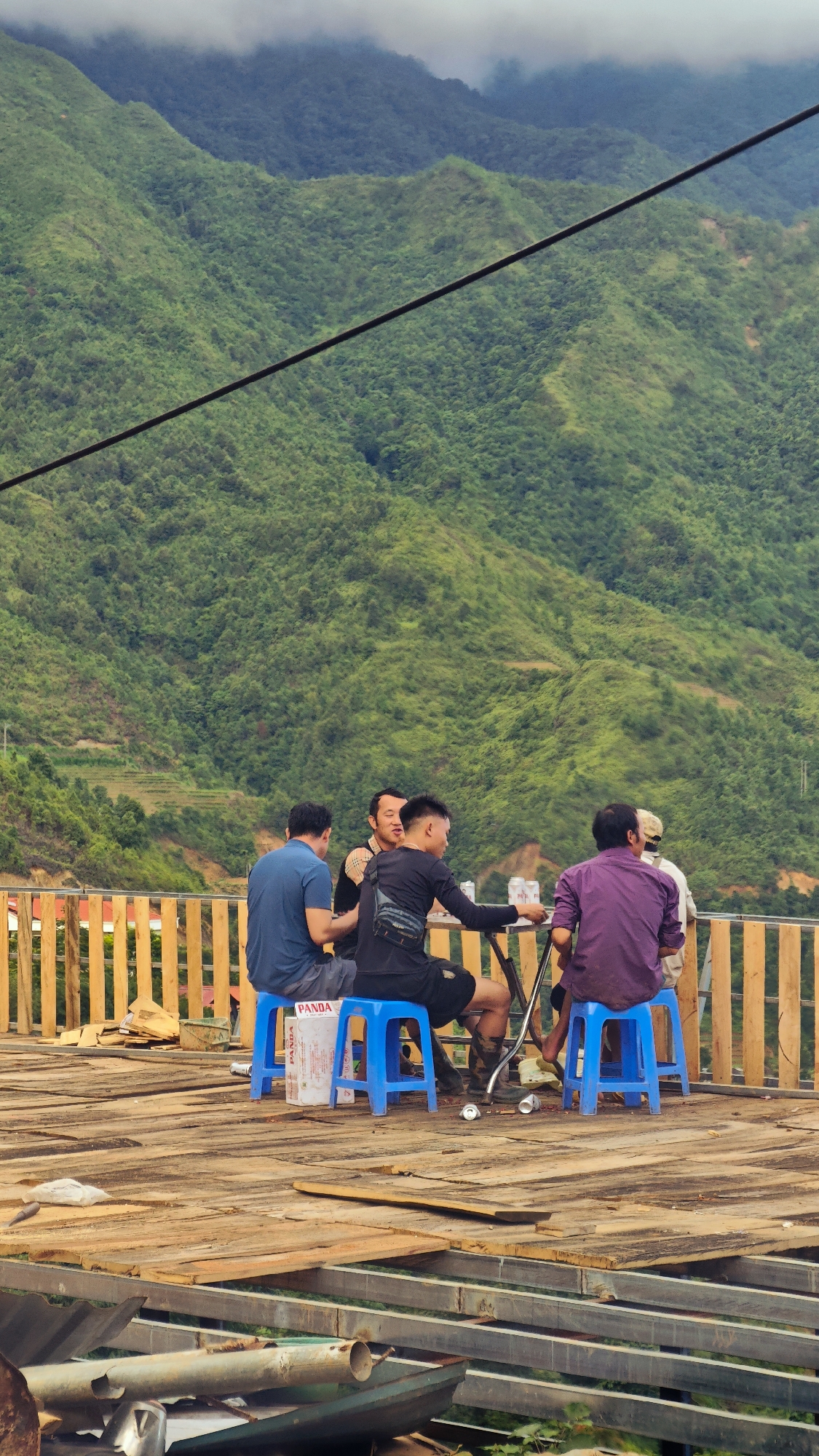 This image depicts five adult males gathered on a wooden deck in Xín Vàng, Vietnam, with a panoramic view of lush green mountains under a cloudy sky. Four men are seated on blue plastic stools around a small circular table with a metal frame. The fifth man is partially visible, seated behind the man in purple, also on a blue stool.
From left to right, the visible individuals are: a man in a blue t-shirt and grey shorts, facing right; a man in a plaid shirt over a dark t-shirt, facing right and looking towards the man to his immediate right; a man in a black long-sleeved shirt and black shorts, wearing a black fanny pack, facing left and looking towards the man in the plaid shirt; and a man in a purple long-sleeved shirt, with his back mostly to the viewer, facing left.
On the table are multiple silver cans, consistent with beer cans, and at least one clear plastic bottle. A white cardboard box with "PANDA" printed on it is positioned between the blue stools of the leftmost and center-right men. The wooden deck is constructed from rough planks, and several empty silver cans are scattered on its surface near the stools. A light brown wooden railing runs along the edge of the deck behind the seated group.
The background features steep, verdant mountains densely covered in trees and vegetation, extending to the horizon. The sky above is overcast with grey clouds. A dark, thick cable or wire stretches diagonally across the upper left to the mid-right portion of the image. In the lower foreground, beneath the wooden deck, various construction materials are visible, including metal beams, corrugated sheets, and metal pipes.