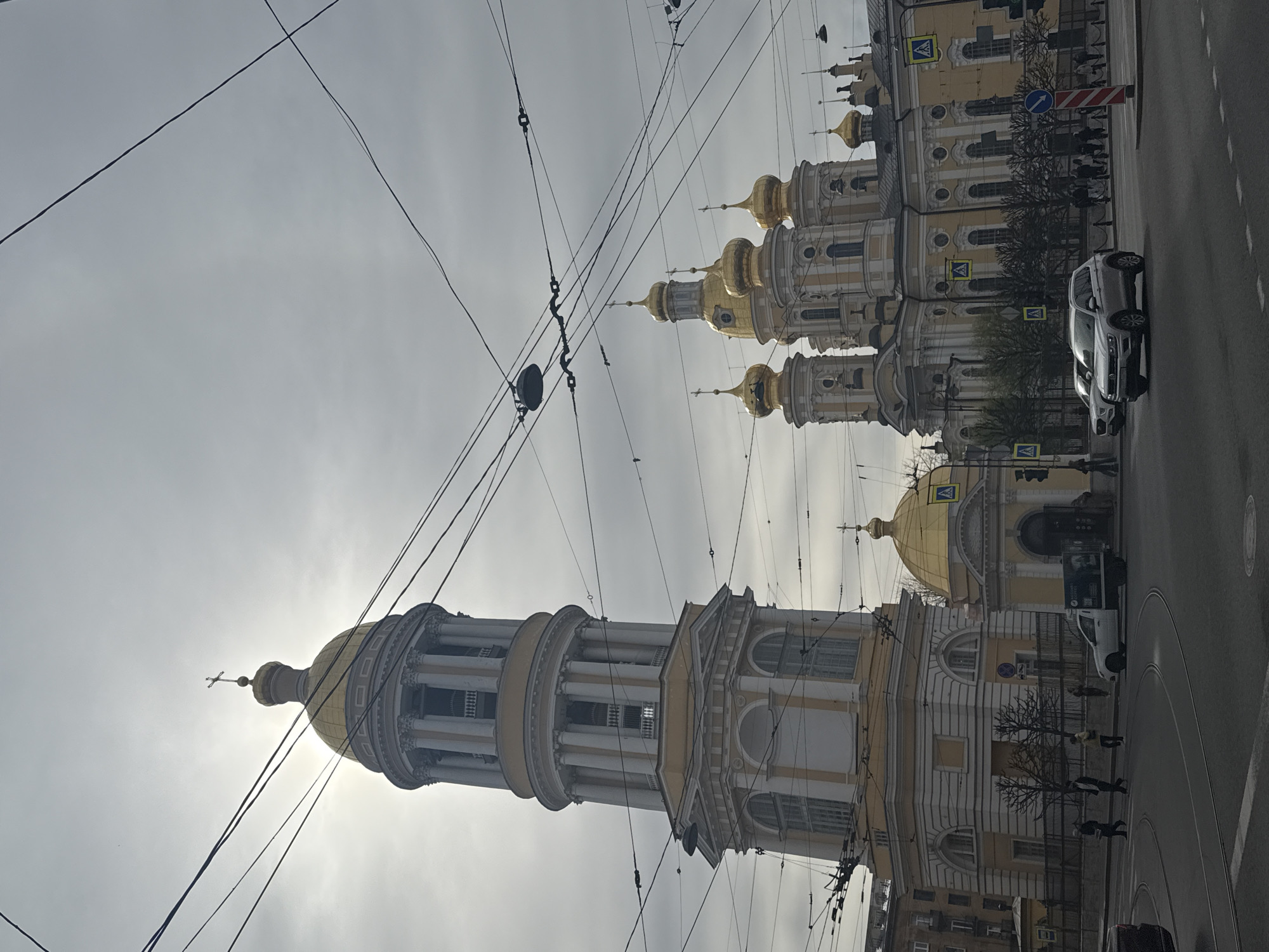 The image presents an urban street scene in Saint Petersburg, Russia, characterized by grand religious architecture and a busy street environment. The photograph is taken from a low angle, looking up at the prominent buildings and an overcast sky.
The central subject is a majestic, multi-tiered bell tower with classical columns, topped by a large golden dome and a cross, positioned on the left side of the frame. The sun is behind its dome, creating a bright silhouette effect. To the right, a complex of buildings, likely part of the same religious ensemble, features multiple smaller golden domes and spires. These structures display ornate yellow or cream facades with white architectural elements. Further to the right, a smaller building with a single golden dome is also visible along the street.
Numerous dark overhead wires crisscross the sky, indicating an urban setting with potential public transport infrastructure. Below the buildings, a wet asphalt road stretches across the lower portion of the frame, showing white lane markings and several pedestrian crossing stripes. A silver SUV is prominently visible on the road, alongside other vehicles and a white van further down. Several pedestrians, dressed in coats, are scattered along the sidewalks near the buildings, suggesting cooler weather. Bare trees, devoid of leaves, line the street, indicating late autumn, winter, or early spring.
The sky is uniformly overcast, contributing to a soft, diffuse light. Notable details include yellow square pedestrian crossing signs and a blue circular sign with a white arrow on the street. No specific text on buildings or vehicles is clearly legible. The scene conveys a sense of a historic city under cool, possibly damp, daytime conditions.