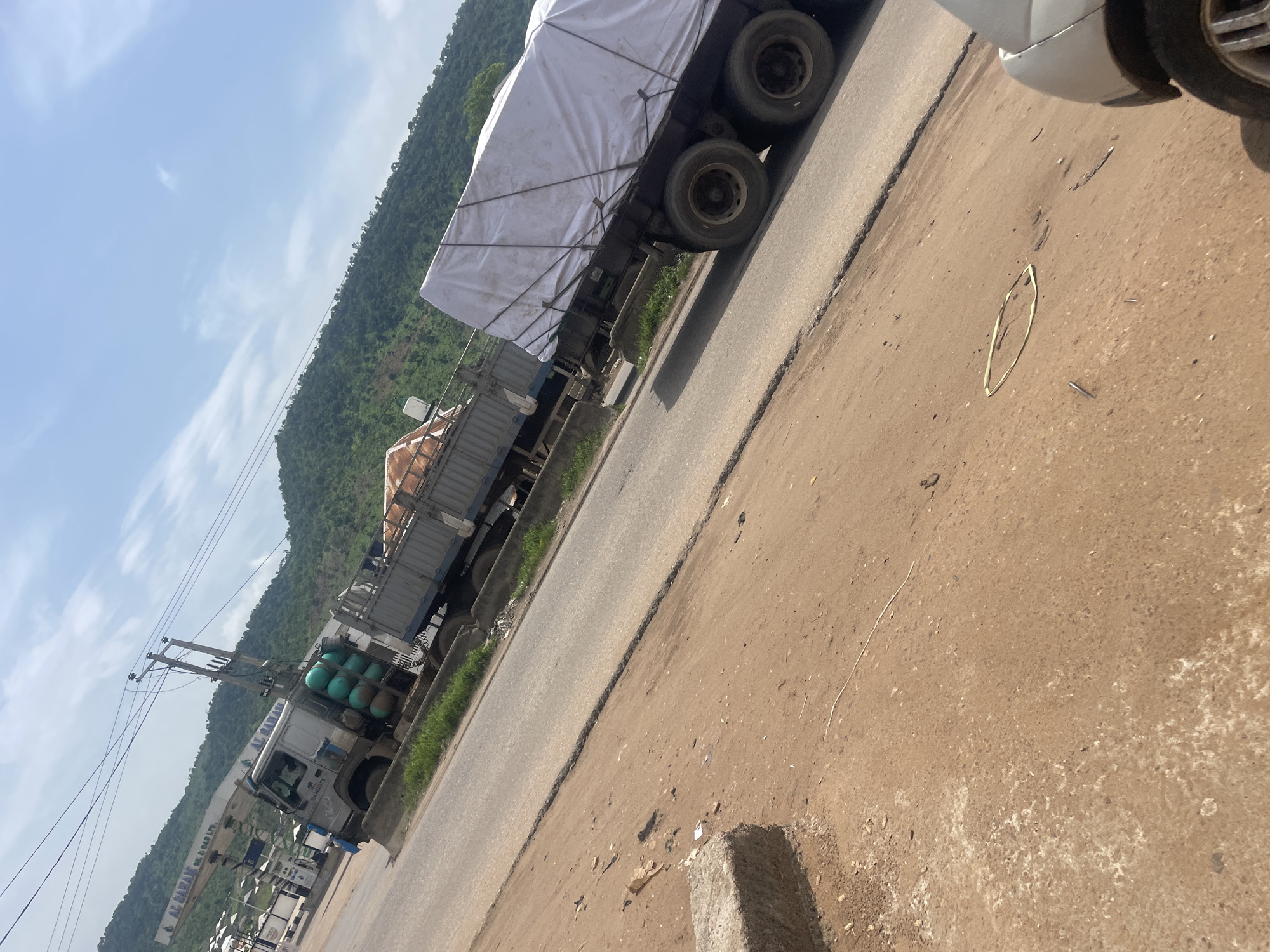 The image depicts a roadside scene in Lokoja, Nigeria, with a focus on two large trucks. The truck in the foreground is a flatbed trailer with its cargo covered by a white tarp, secured with ropes. The truck's tires are visible, and it is positioned on a paved road. In the background, another truck is partially visible, parked near a fuel station with a prominent "AL-SAMAN OIL & GAS LTD." sign. This truck appears to be a tanker, indicated by several large green cylindrical tanks mounted on its side. The surrounding environment includes a dirt embankment on the left, a paved road, and a backdrop of lush green hills under a partly cloudy sky. The overall impression is of a commercial area along a major transportation route.