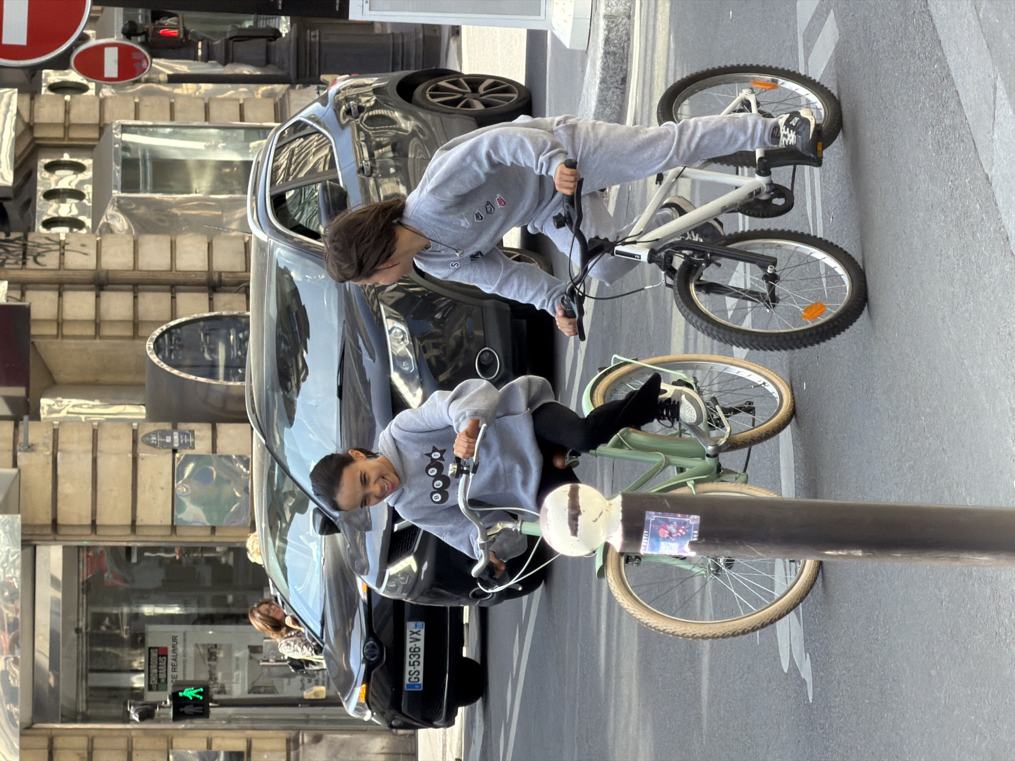 Two young people are captured on a bright, sunny day on a city street, likely in Paris, France. The image is taken from a slightly elevated perspective, looking down at the street level.
In the foreground, a person with dark hair, wearing a grey hoodie and sweatpants, stands on the pedals of a white bicycle. They are looking forward, with their body angled over the handlebars. Behind them and to the right, another person with dark hair, also in a grey hoodie and black pants, sits on a light green bicycle. This individual is smiling broadly and looking towards the camera. The light green bike has distinctive light brown or tan-colored tires. Both bikes are modern mountain bike or urban hybrid styles.
Behind the cyclists, a black car is parked, with its license plate "GS-536-VX" clearly visible, indicating a French registration. Reflections of the surrounding buildings and sky are visible on the car's windows and body. The background features typical Parisian architecture, with multi-story buildings and reflections of storefronts. Visible text on a storefront includes "FOURRURES CHARLES" and "CE REAUMUR," referencing a street in Paris. Red and white traffic signs (likely "no entry") are seen near the top left. A pedestrian crossing signal shows a green "walk" symbol on a pole in the middle distance. A dark street pole with a small colorful sticker is present in the lower right, partially obscuring the green bicycle. The street itself has painted white lines.