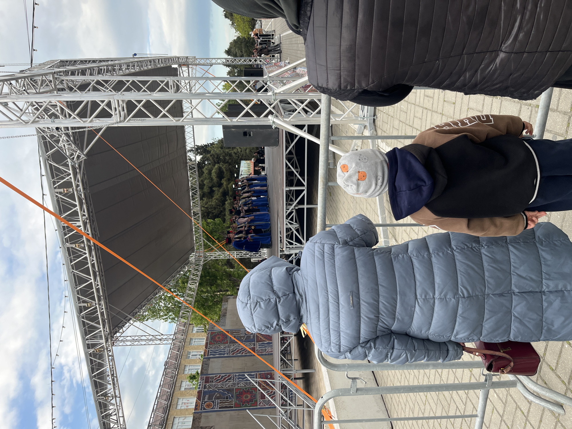 The image captures an outdoor scene in Kaspiysk, Russia, featuring a temporary stage structure with a gray cover. A group of performers in blue outfits is visible on the stage. In the foreground, there are people watching the performance, including a person in a light blue puffer jacket and a child in a brown and black hoodie with a white hat with bear prints. A metal barricade separates the audience from the stage area. To the side of the stage, there is a building with a large mural on its facade. Orange straps are connected to the stage structure. The sky is cloudy.
