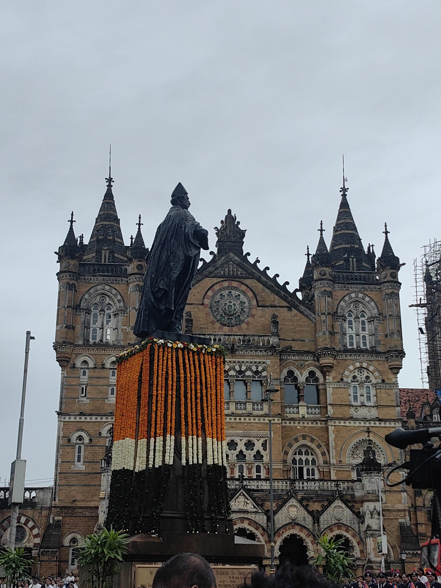 This is a photograph of the Chhatrapati Shivaji Maharaj Terminus (CSMT) in Mumbai, India, formerly known as Victoria Terminus. A large statue of a man in a cape and hat stands on a decorated pedestal in front of the historic building. The pedestal is adorned with garlands of marigolds and green leaves, arranged in a pattern reminiscent of the Indian flag. The CSMT building itself is a UNESCO World Heritage Site, characterized by its Gothic Revival architecture, with spires, arched windows, and intricate stone carvings. The sky is overcast, suggesting a cloudy day. Several people are visible in the foreground, some appearing to be part of a crowd. A camera on a tripod is also present, indicating that the scene is likely being documented.