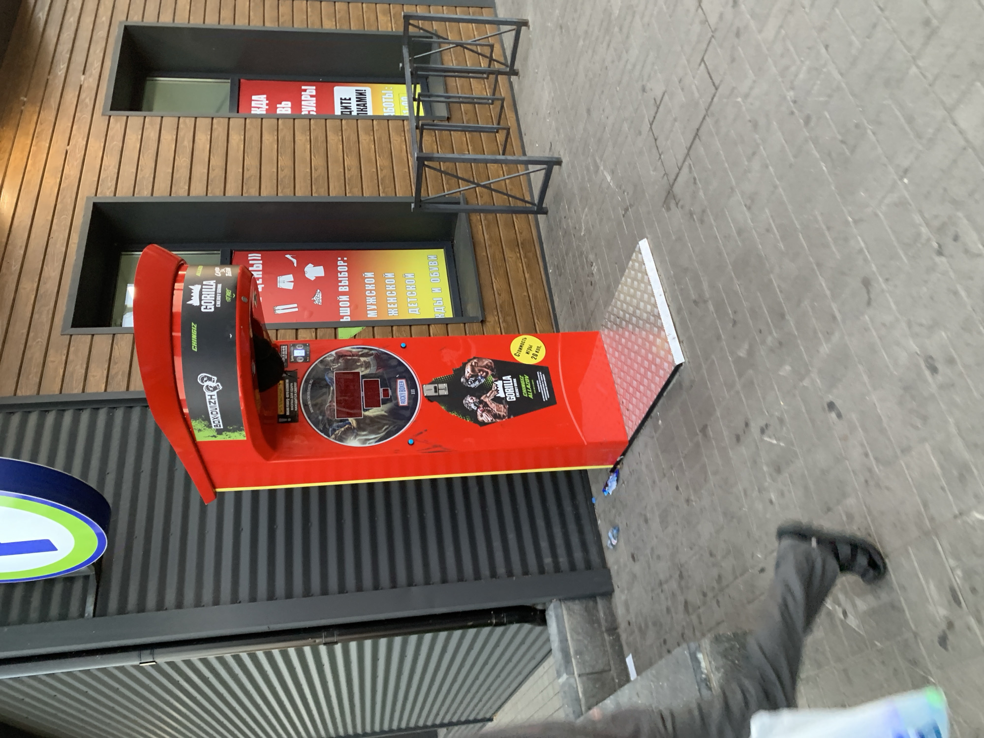 In Kapyĺ, Belarus, a red boxing machine, branded with "GORILLA ENERGY DRINK" and "ROCKY BOXER," stands on a grey tiled sidewalk. The machine features a digital display, images of muscular figures, and text advertising clothing and footwear choices. To the left of the machine, a person wearing grey pants and dark sandals walks away from the camera, carrying a white bag. The background consists of a building with a dark corrugated metal facade and a wooden paneled section with two dark-framed windows. A metal bicycle rack is visible to the right of the machine. Debris, including plastic bottles, litters the sidewalk around the machine.
