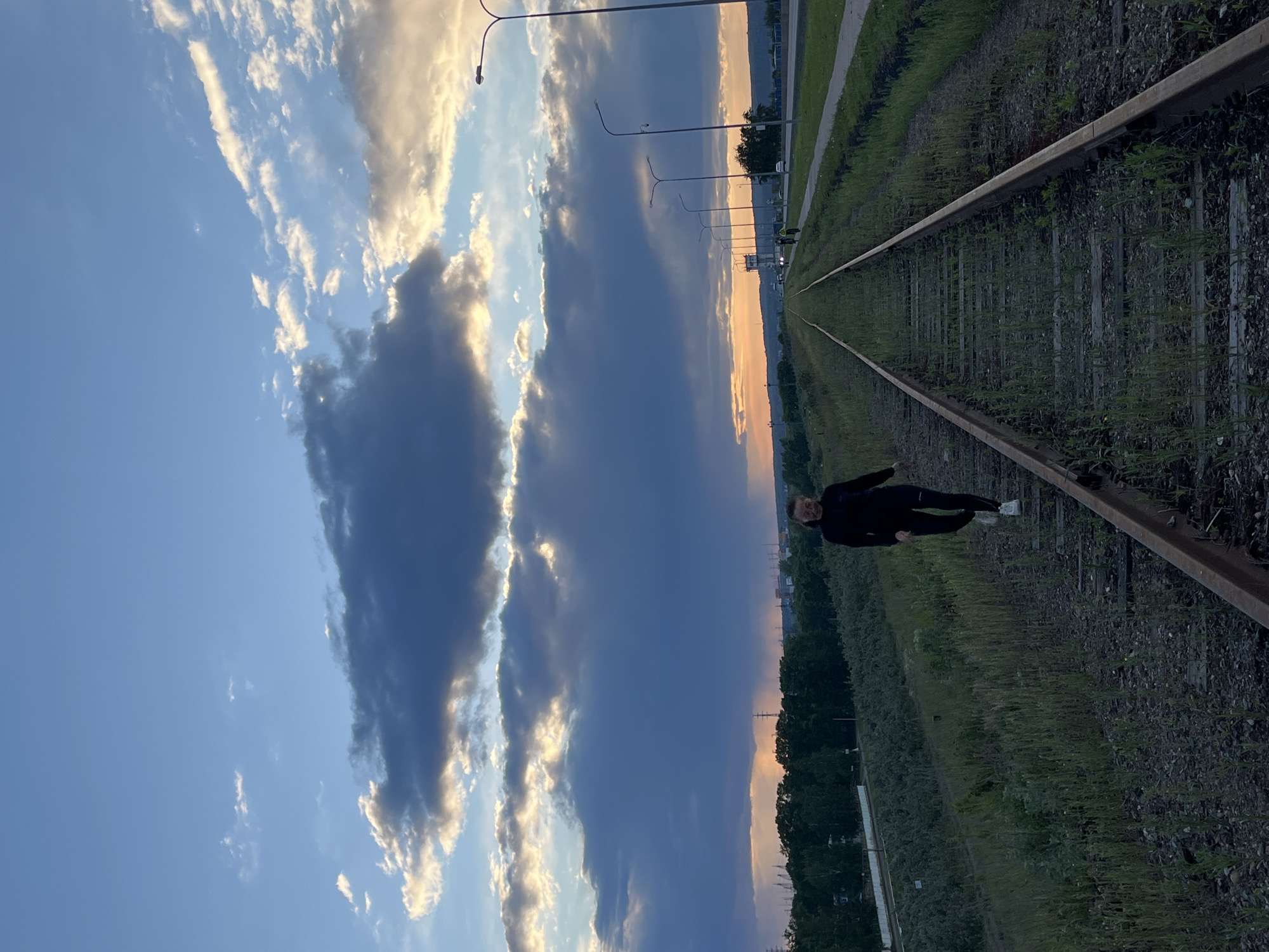The image shows a person balancing on a railroad track in Chaikovsky, Russia. The sky is filled with dramatic clouds at sunset. Streetlights line a road parallel to the tracks. Vegetation grows densely alongside the railway. The person is dressed in black and wearing white shoes, facing toward the end of the tracks.